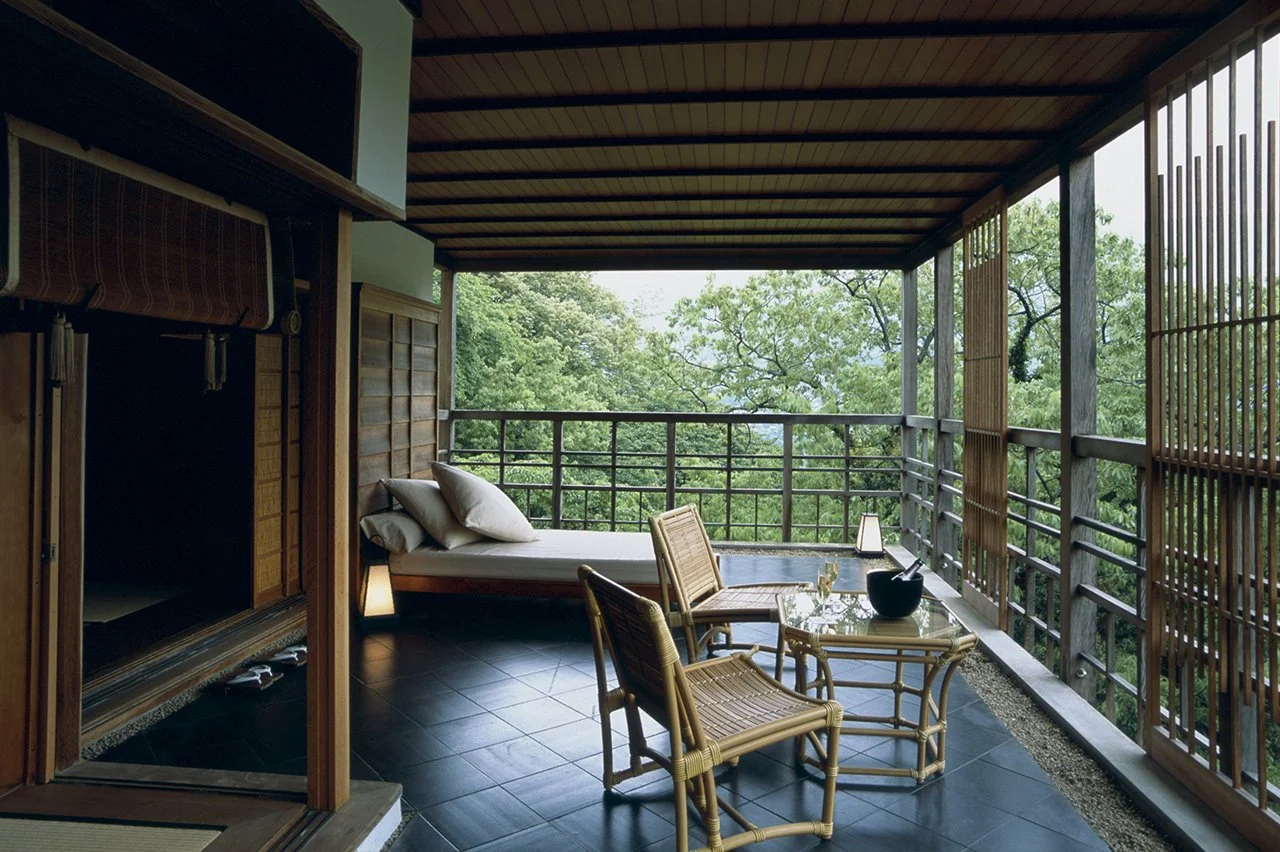 Balcony with a bed, bamboo chairs, and a glass table overlooking green trees.