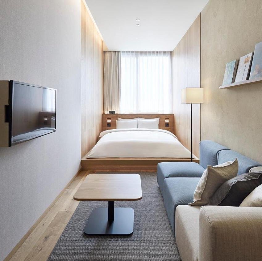 Tourist hotel room with a bed, a blue sofa, a white armchair, a floor lamp, a wall-mounted TV, and window with curtains.