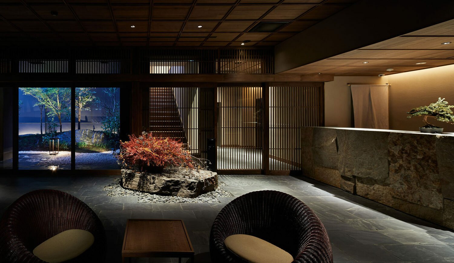 Interior of a modern building with a Japanese-inspired design, featuring a rock garden with a red-leafed plant, wooden slatted walls, dark tiled flooring, and seating area with two woven chairs and a small wooden table, illuminated by soft lighting.