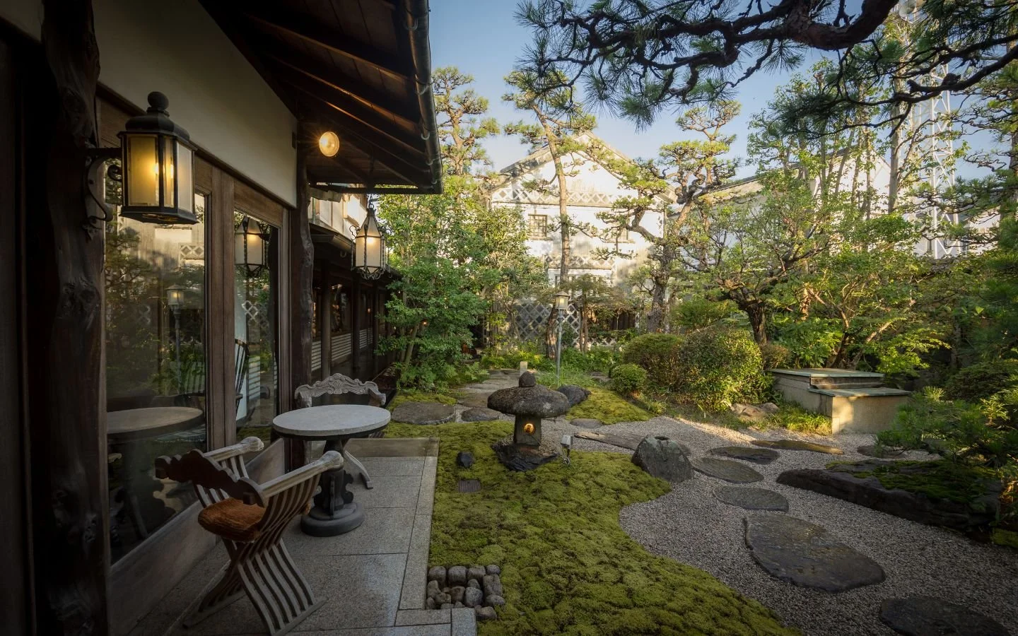 A peaceful Japanese-style garden with a stone lantern, stepping stones, lush green trees, and shrubs adjacent to a traditional wooden building with outdoor seating and lanterns.