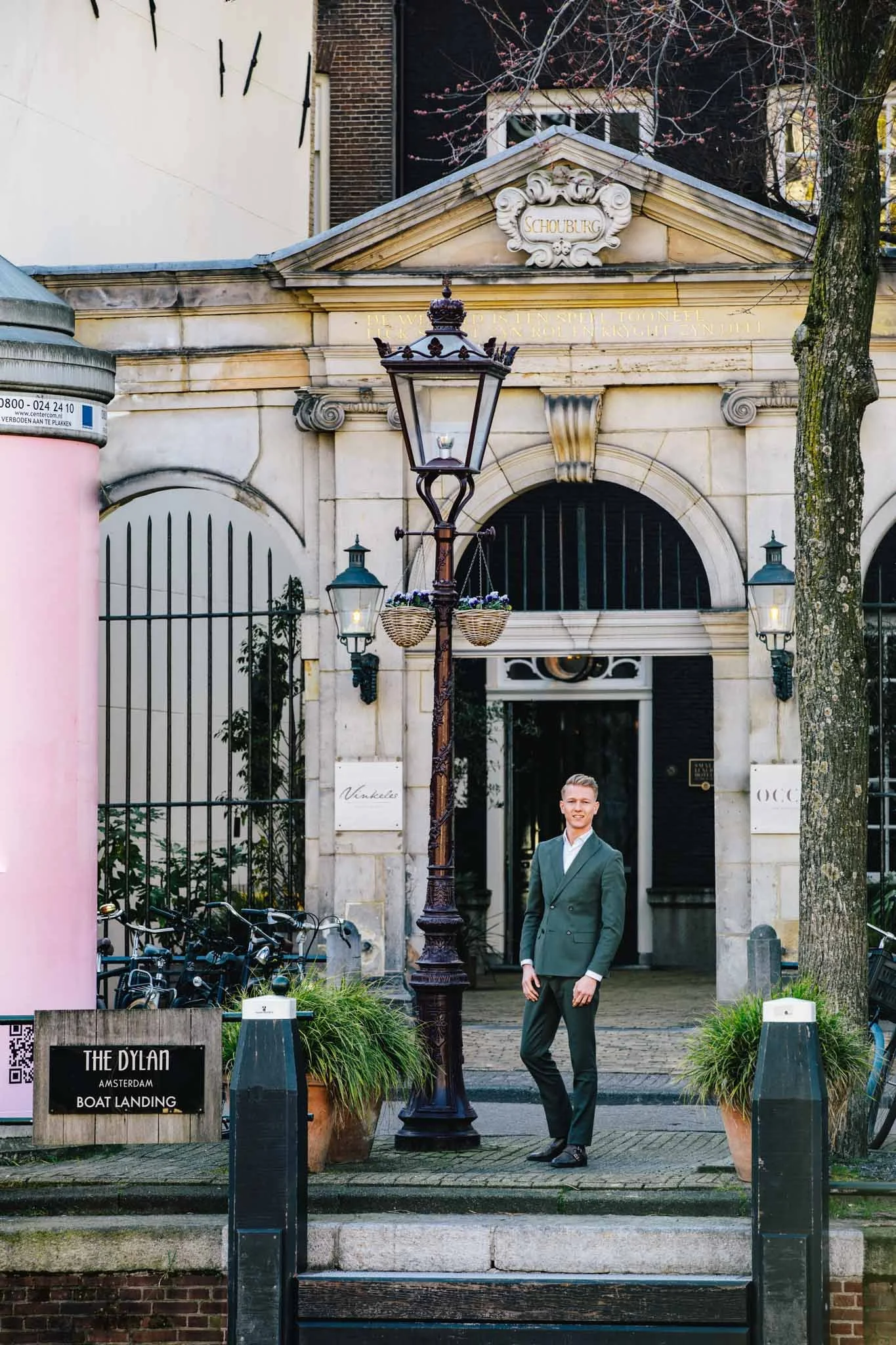 A man in a green suit smiling standing on a sidewalk in front of an ornate building in Amsterdam. There are bicycles and a sign reading 'The Dylan Amsterdam Boat Landing' nearby. A vintage street lamp is also in the scene.