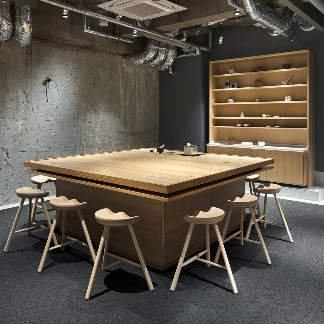 A modern, minimalist kitchen with a large wooden table surrounded by eight matching stools, an open wooden shelf with various dishes, and exposed industrial-style ductwork on the ceiling.