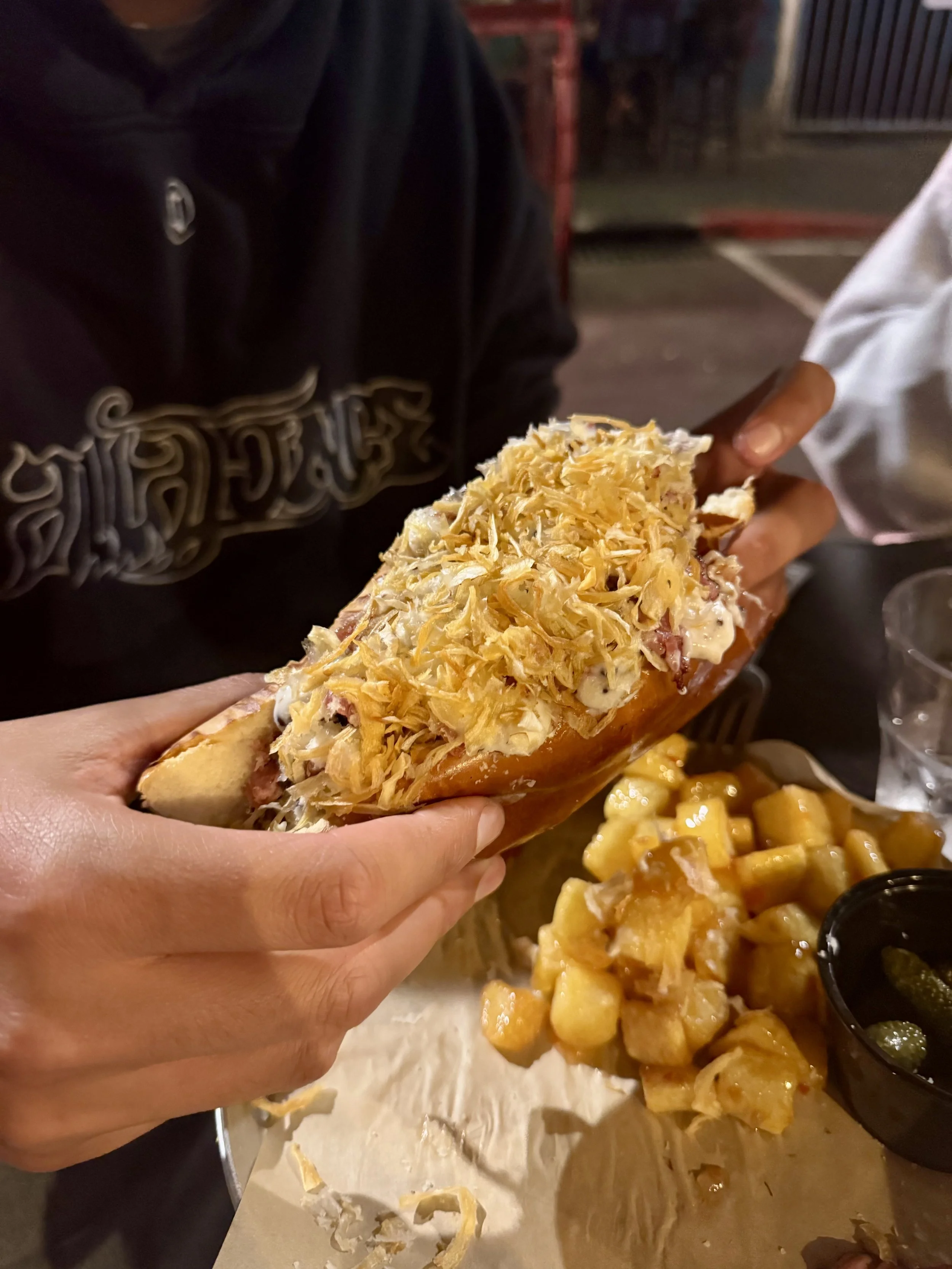 Person holding a sandwich topped with shredded cheese, with cubed potatoes and a cup of pickles on the table.