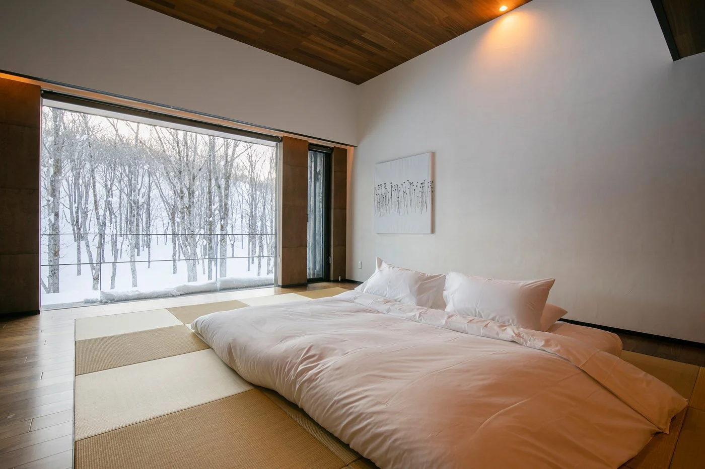 Minimalist bedroom with a large bed, white bedding, and pillows, featuring a floor-to-ceiling window with a view of snowy trees outside, and a wooden ceiling.