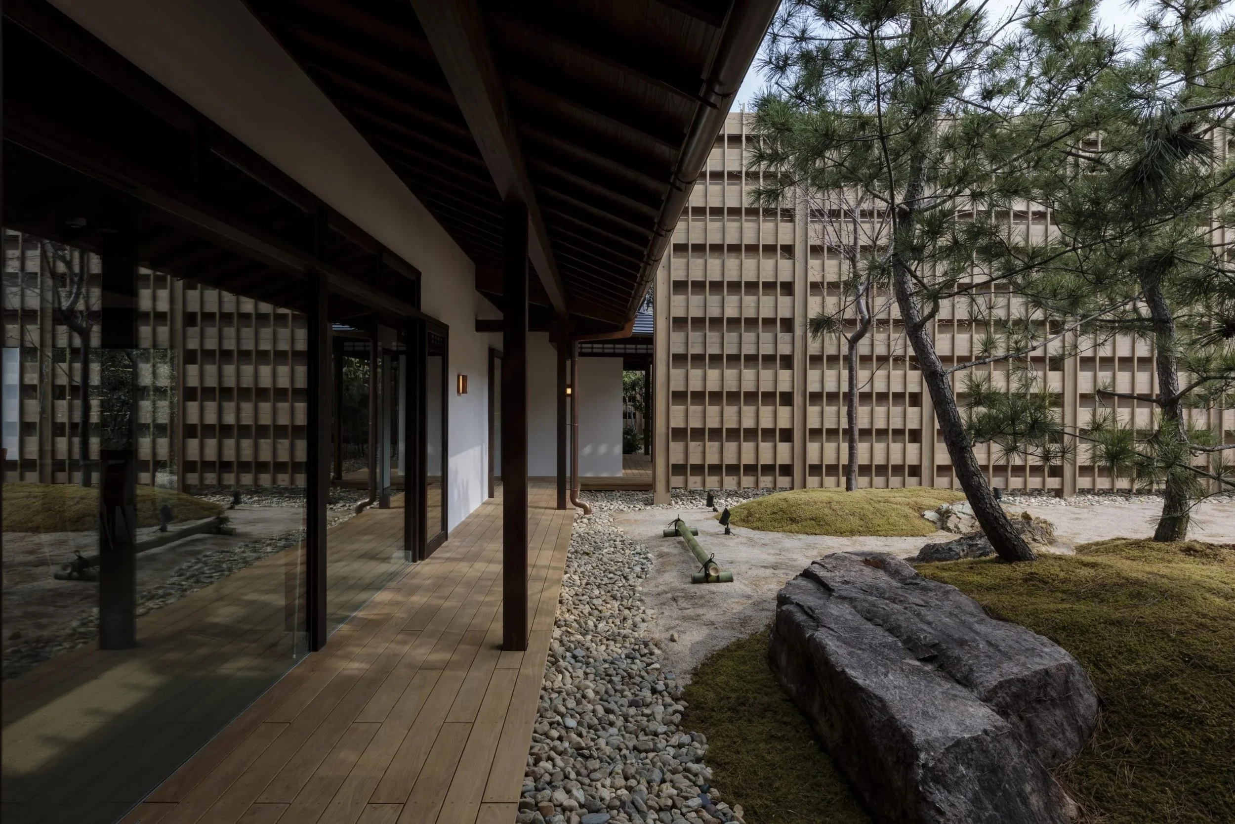 A traditional Japanese-style garden with a wooden walkway, moss-covered mounds, rocks, and a few pine trees, enclosed by a wooden lattice fence, with a building featuring sliding glass doors and wooden beams.