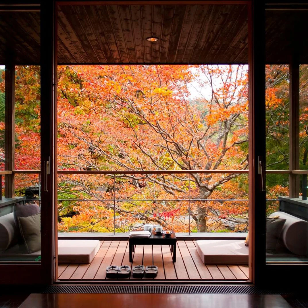 A cozy balcony with cushions and a small table, overlooking colorful autumn trees with orange and yellow leaves.