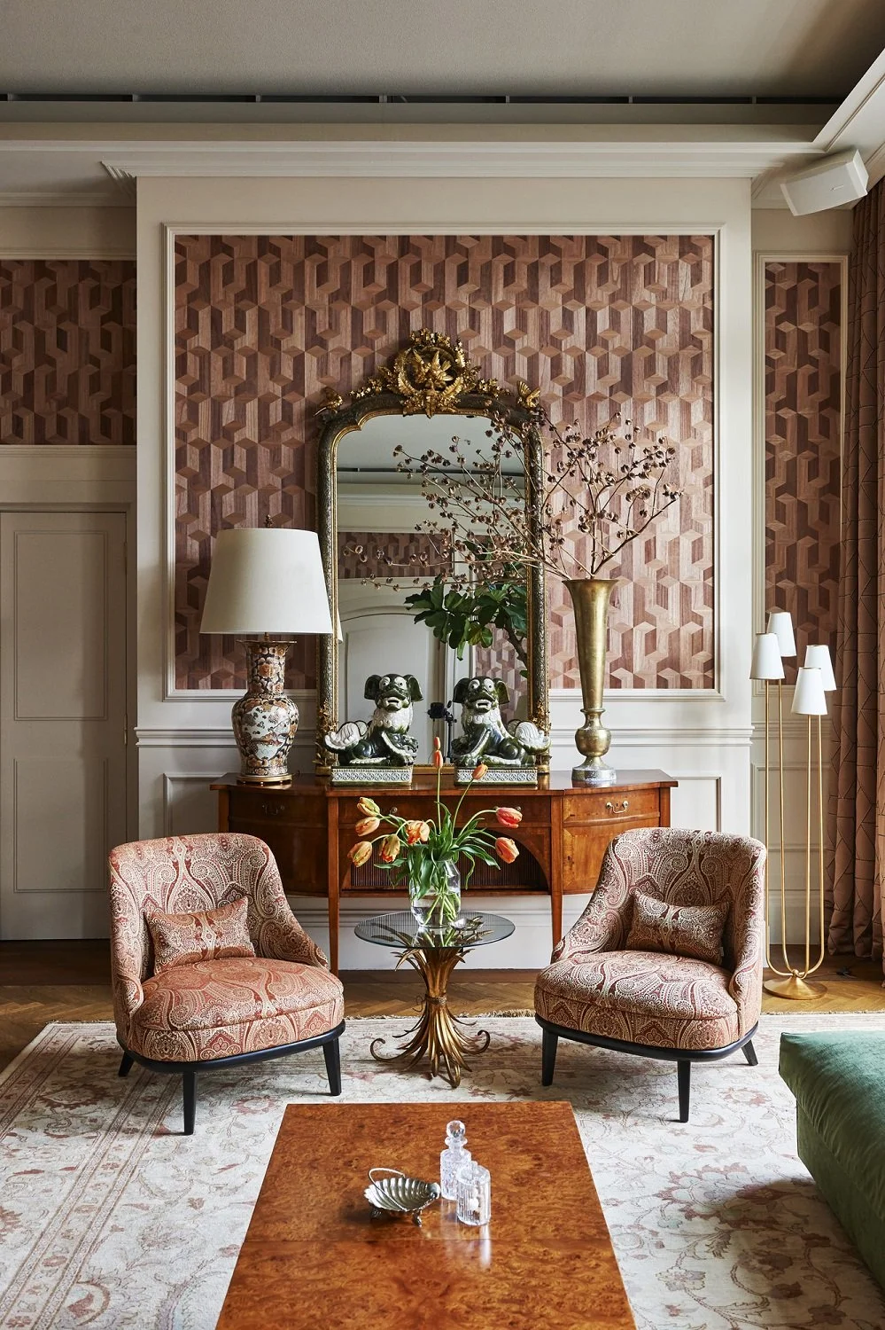 A living room with pink and beige patterned walls, two patterned armchairs with matching pillows, a decorative wooden sideboard with a large ornate mirror, a tall gold vase with branches, a table with a glass top and floral arrangement, other decor including ceramic dogs and lamps, and a cream-colored rug with a border.