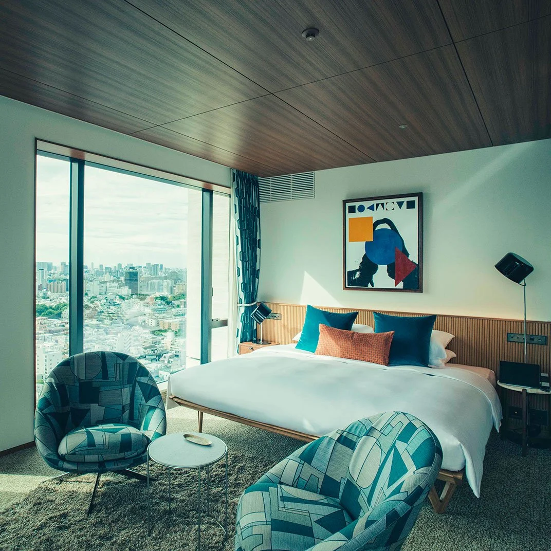 Hotel room with a large bed, two patterned chairs, a small round table, large window with city view, colorful artwork above the bed, and modern decor.