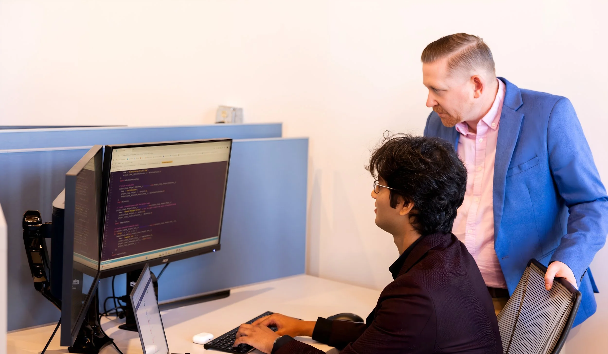 Two men in an office, one sitting at a computer and coding, the other standing and looking at the screen.