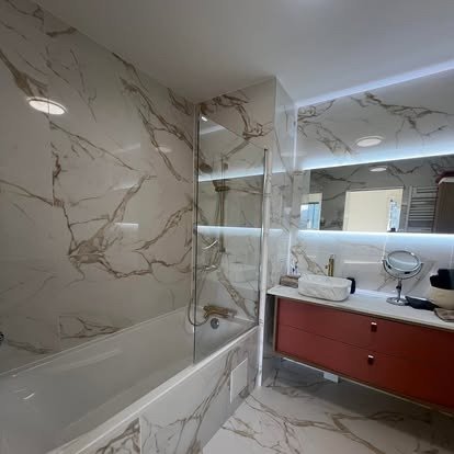 Modern bathroom with marble walls and floor, a bathtub with a glass shower door, a wall-mounted mirror with built-in lighting, and a red vanity cabinet with a countertop mirror.