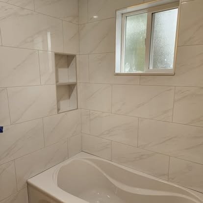 Bathroom with beige tile walls, a built-in corner shelf, a small window, and a white bathtub.