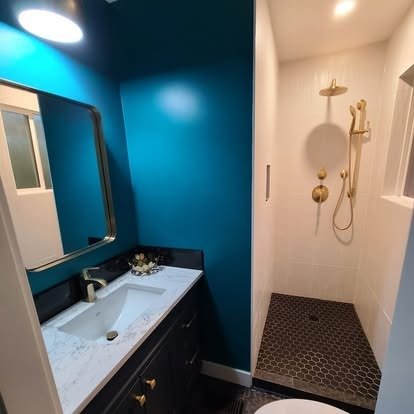 Bathroom with a dark vanity, white marble countertop, and a mirror on a teal wall; adjacent walk-in shower with gold fixtures and a dark hexagonal tile floor.