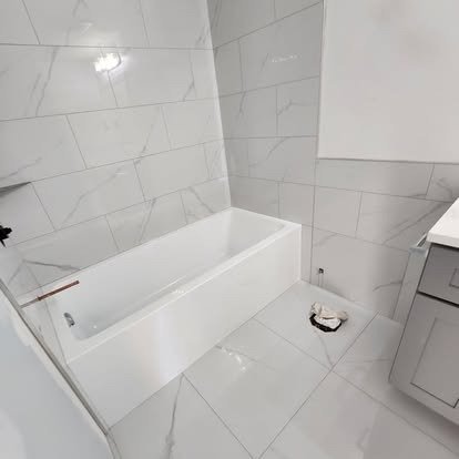Bathroom with white tiled walls and floor, white bathtub, gray vanity, and a small pile of construction debris on the floor.