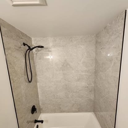 Empty bathroom shower with gray tiled walls, black showerhead, and white bathtub.