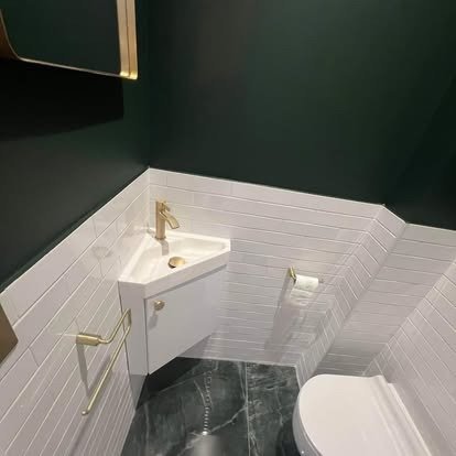 Small bathroom with dark green walls, a white wall-mounted sink with a gold faucet, a gold towel bar, and a white toilet, with white tiling on the lower half of the walls and dark tile flooring.