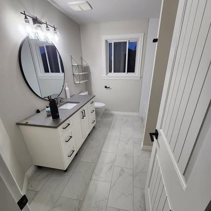 Modern bathroom with white vanity, round mirror, wall-mounted light fixture, toilet, and window, with white walls and marble floor tiles.