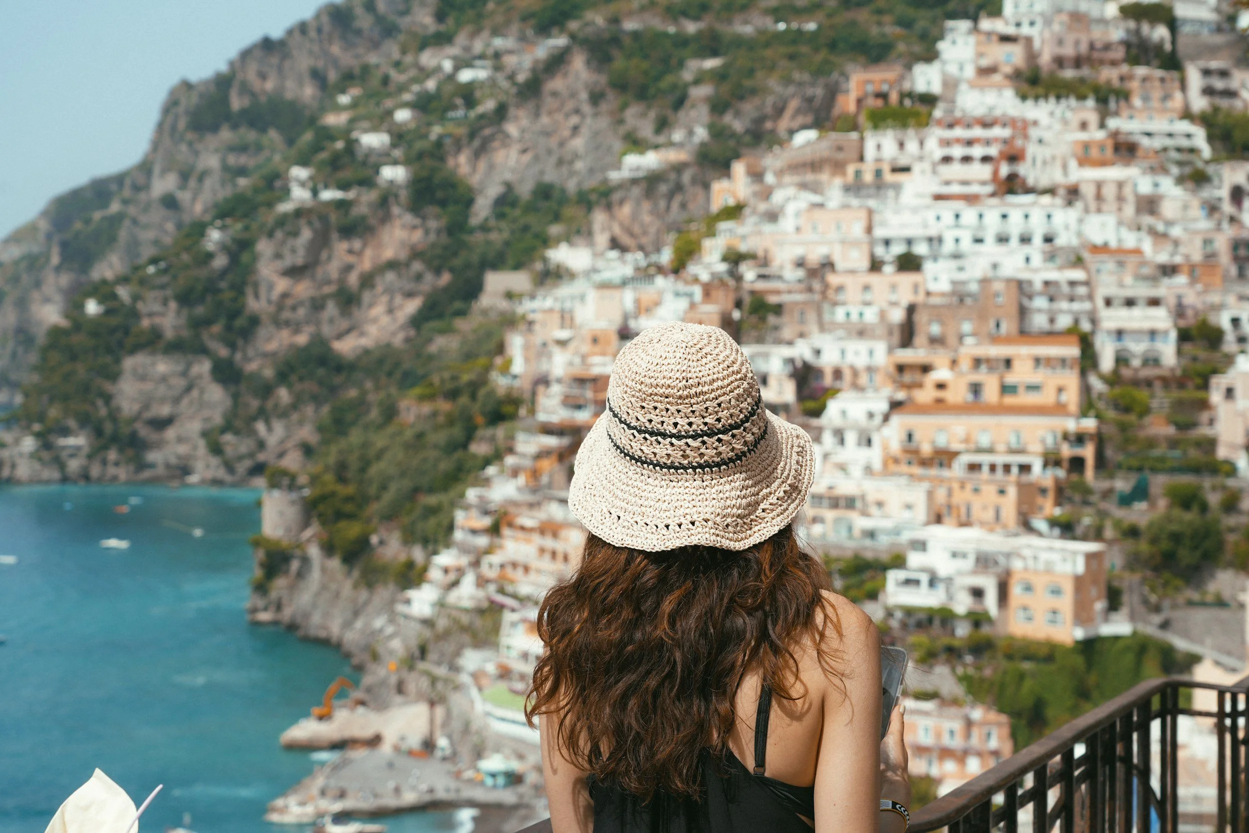 A woman with long curly hair wearing a wide-brimmed straw hat and black sleeveless top standing on a balcony, overlooking a coastal town built on steep cliffs with colorful buildings and a harbor.