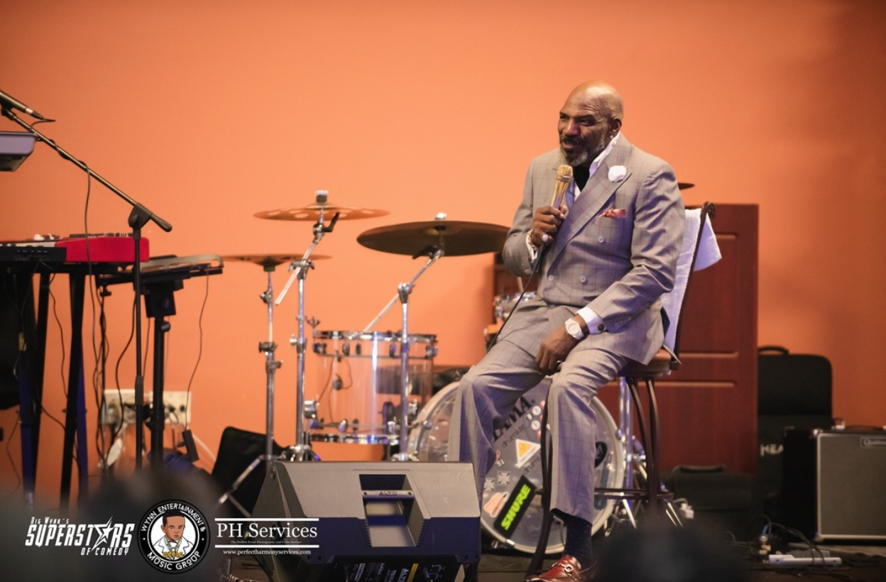 A man in a gray plaid suit sitting on a stool, holding a gold microphone, smiling, with drums and music equipment in the background, on a stage with orange wall.