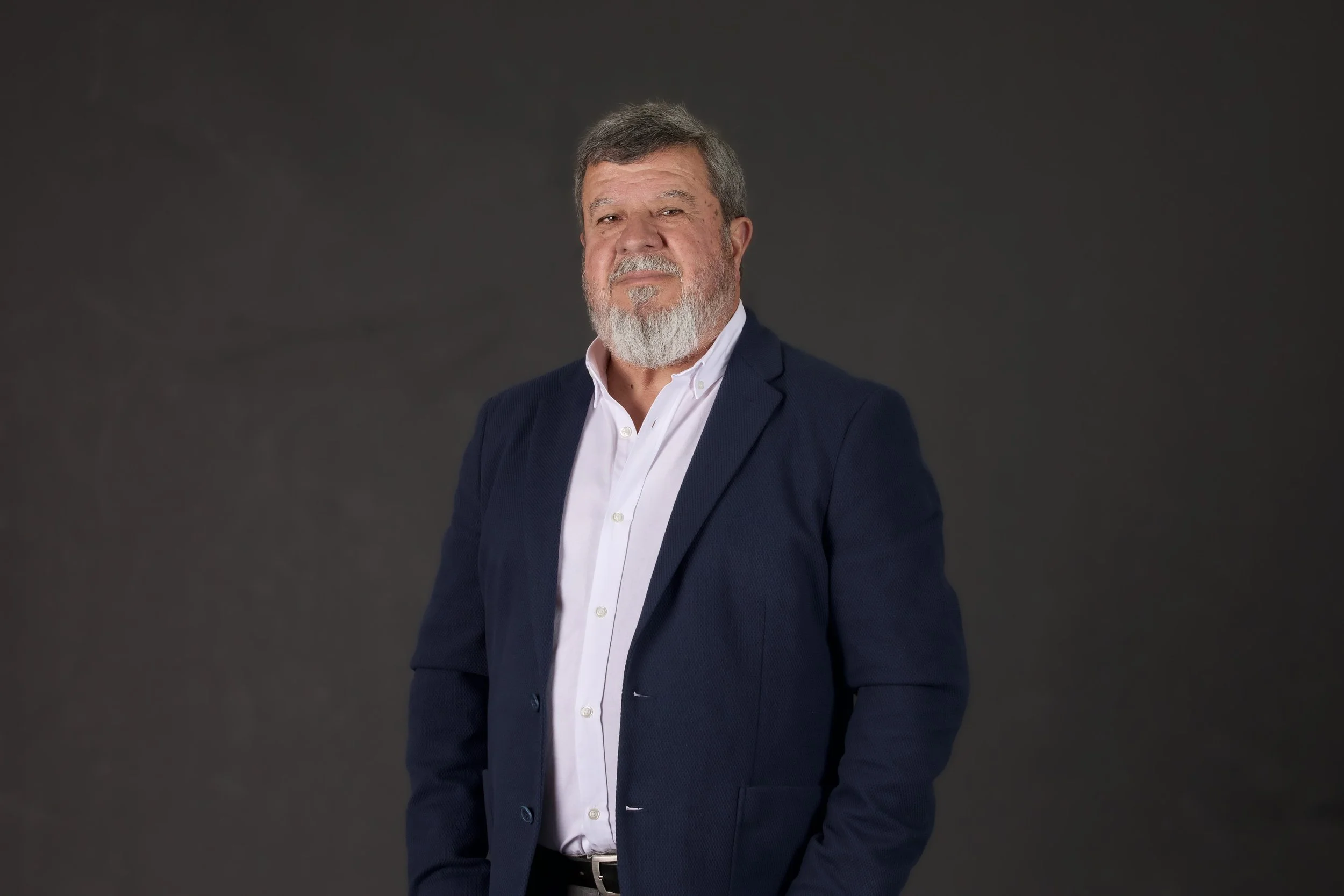 Homem de cabelos grisalhos e barba, usando um terno azul escuro e uma camisa branca, posando em frente a fundo cinza