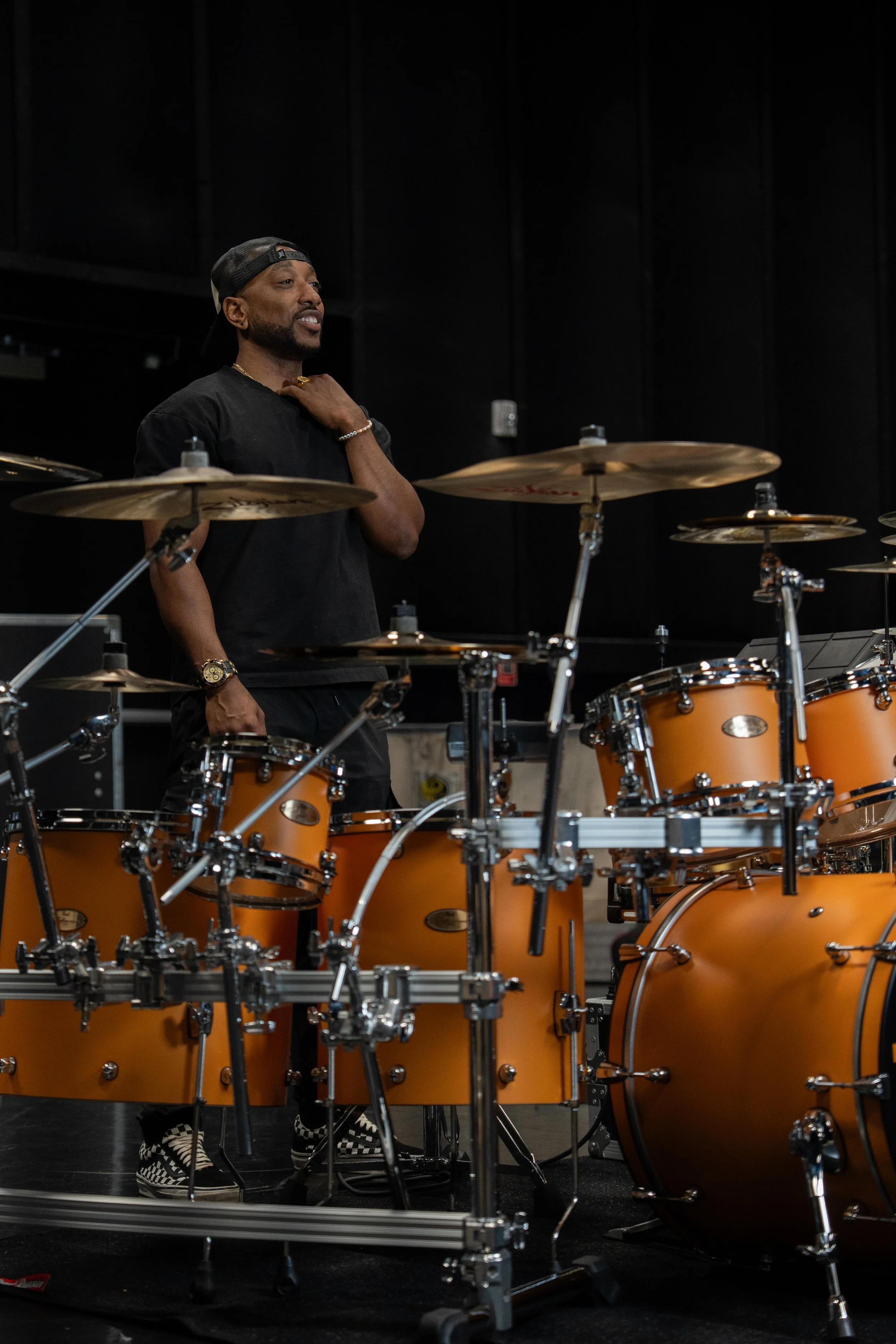 Man standing behind a drum set in a music studio, wearing a black t-shirt and a backward baseball cap.