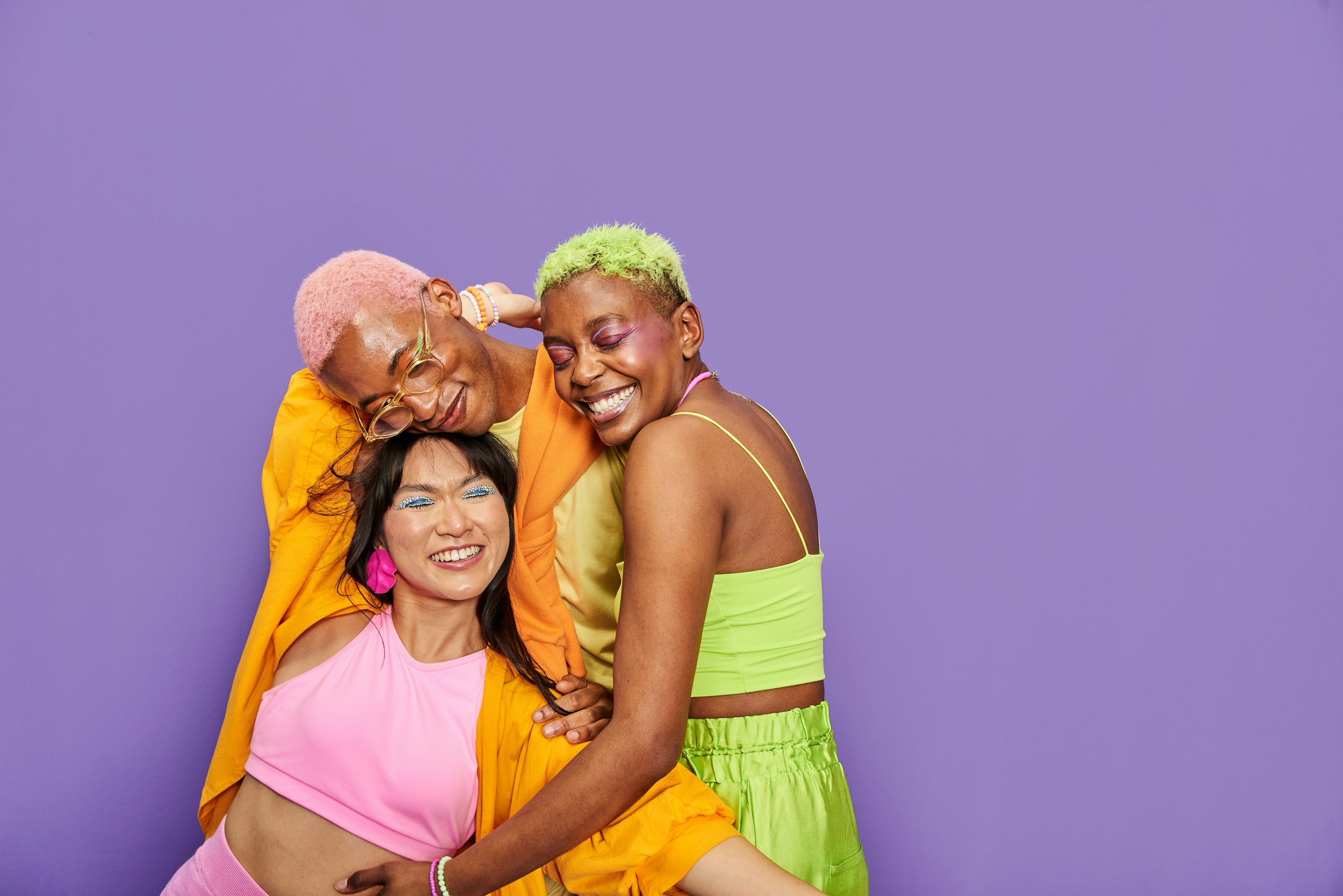 Three ethnically diverse people dressed in colorful, vibrant clothing smiling and hugging against a purple background.