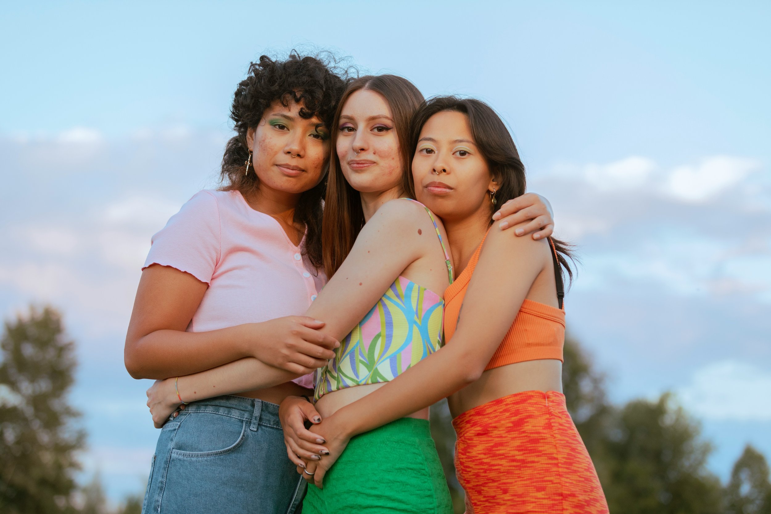 Three women are standing closely together outdoors, embracing, with a blue sky and some trees in the background.