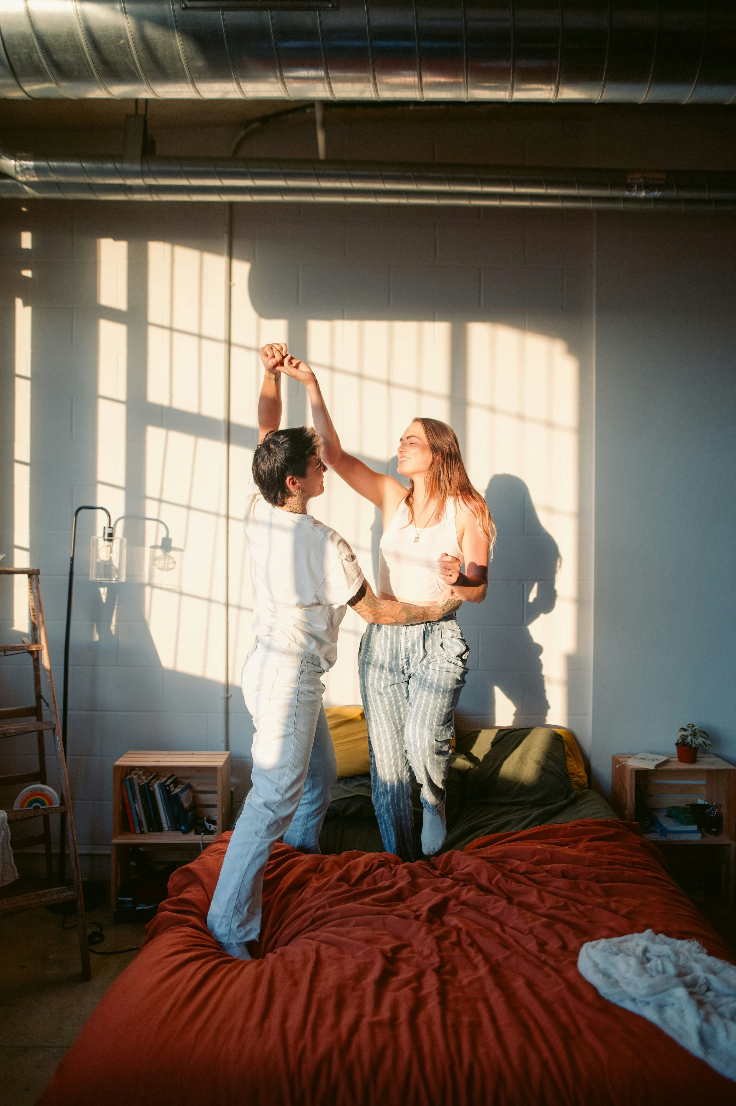 Two women are dancing and holding hands on a bed in a sunlit room, smiling and enjoying the moment.