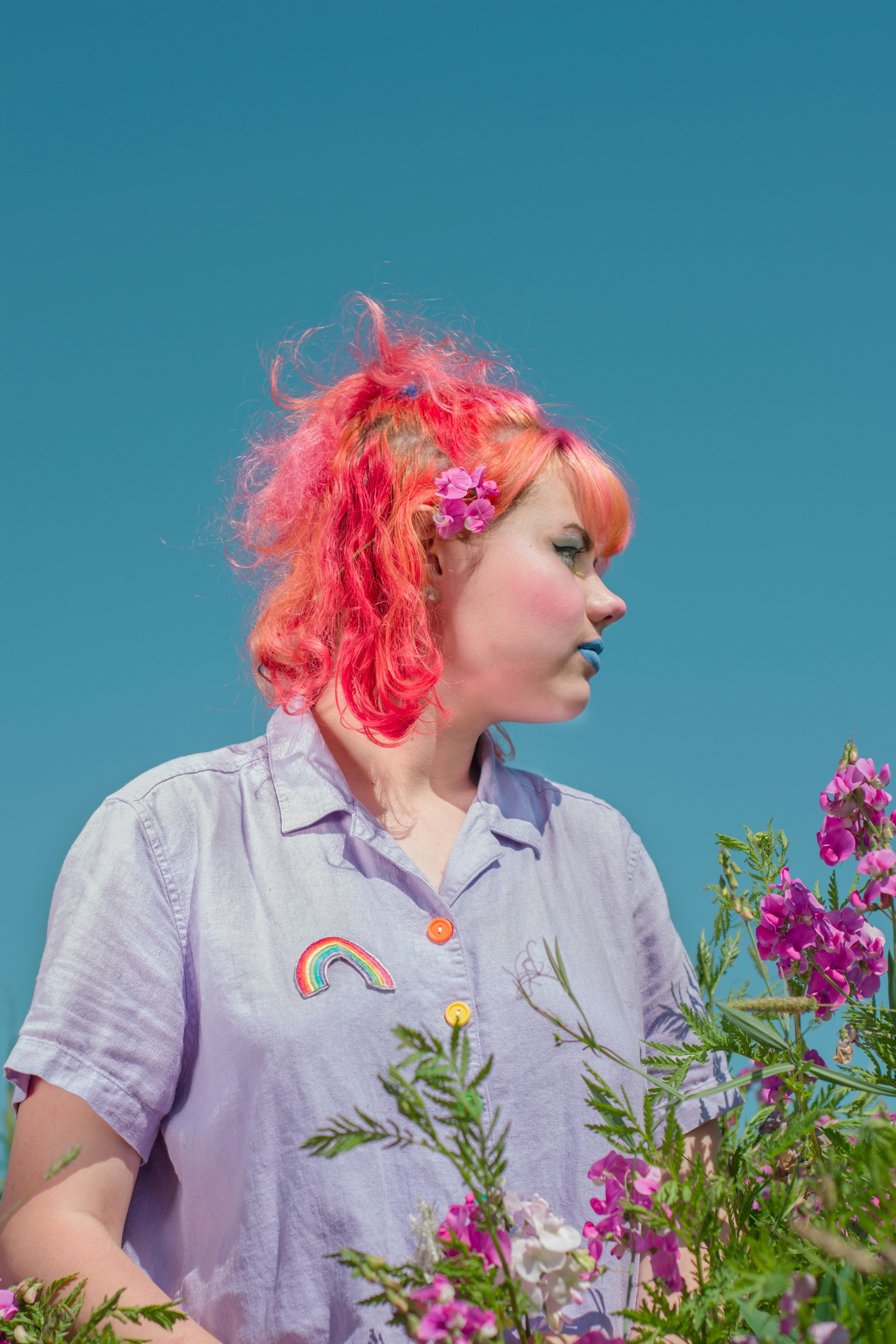 A woman with pink and red curly hair wearing a lavender shirt with rainbow and flower patches, standing among pink and purple flowers against a clear blue sky.