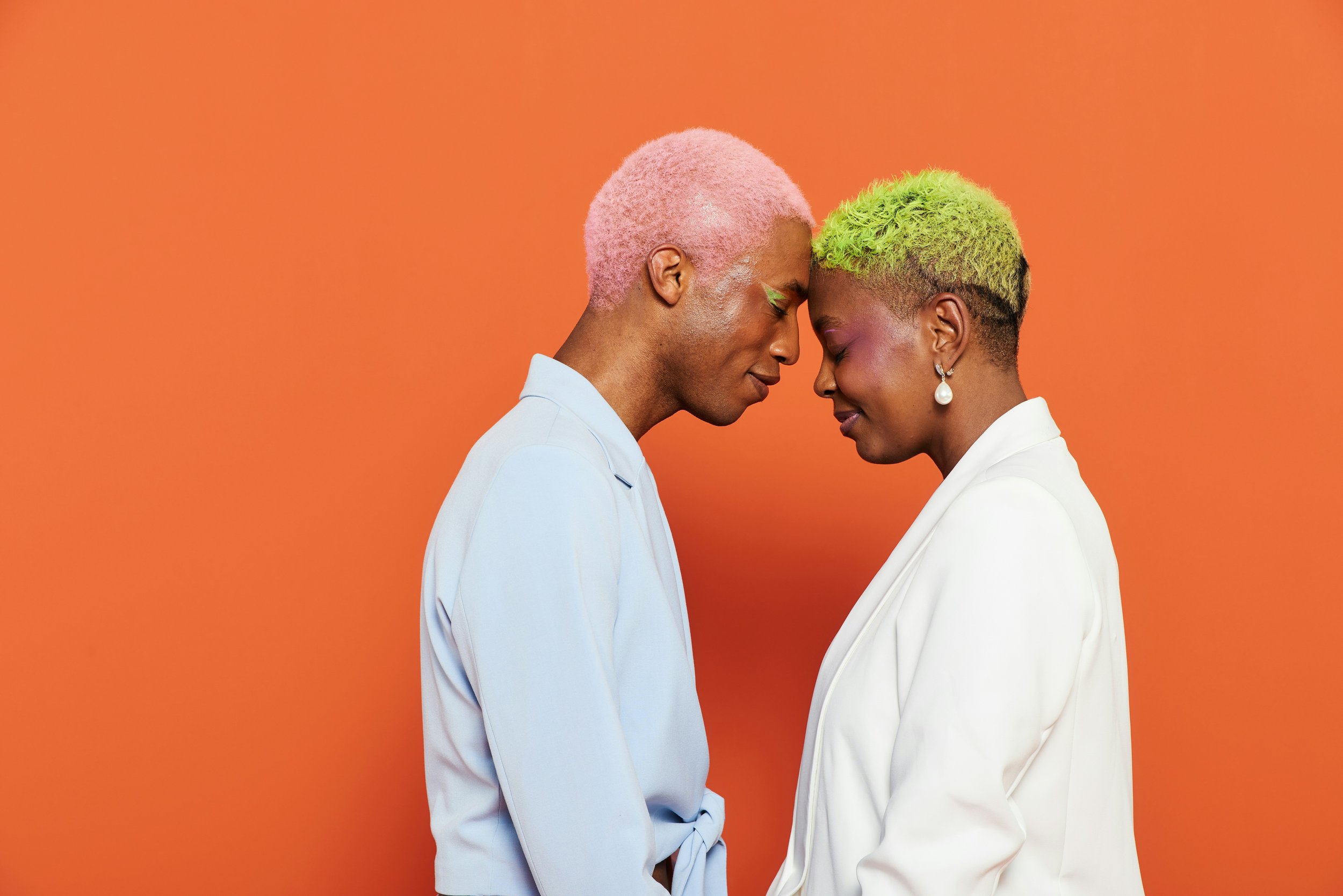 Two women with colorful hair, one with pink hair and the other with green hair, standing close with foreheads touching against an orange background.