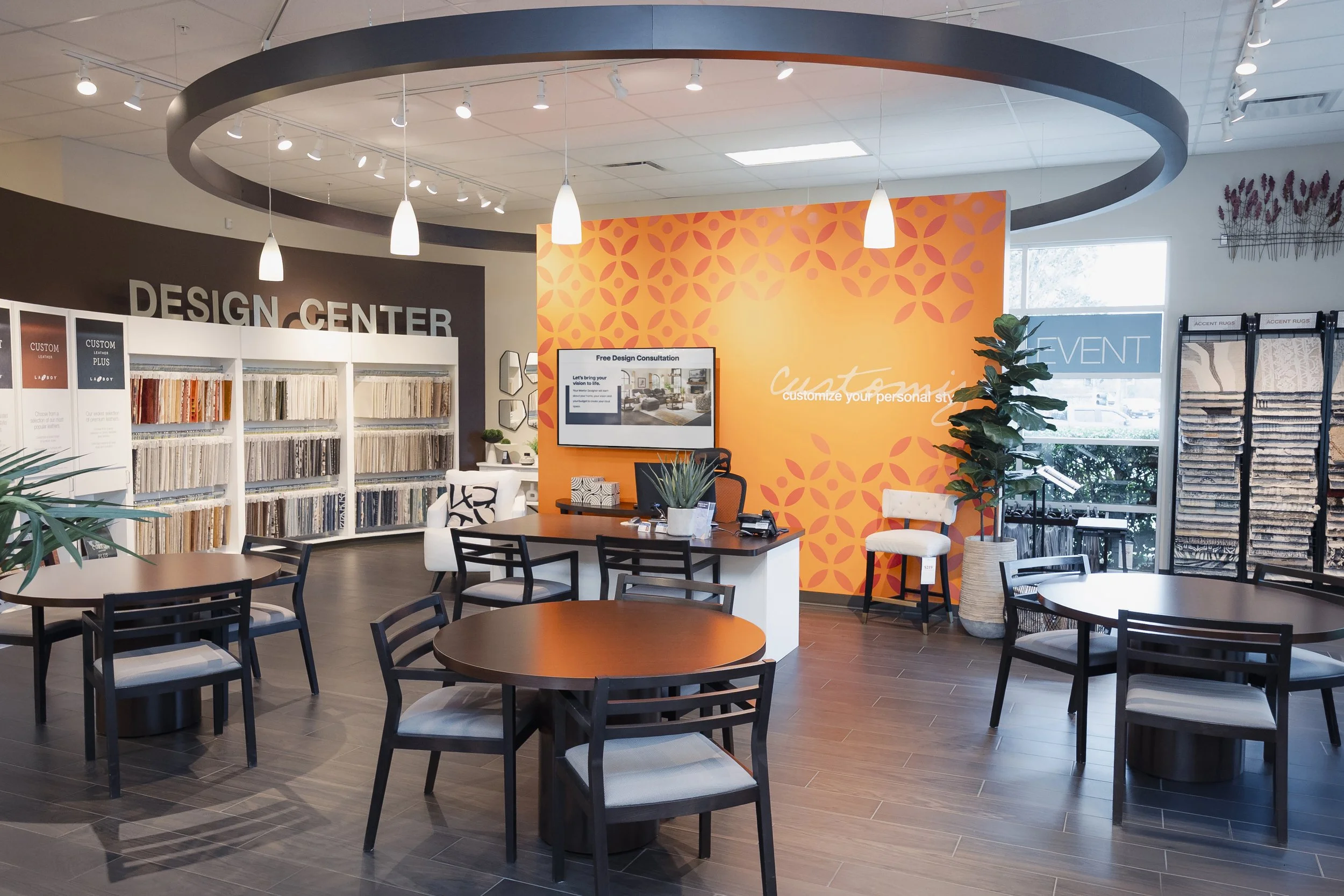 Interior of a design center with round tables and chairs, display shelves with fabric swatches, an orange accent wall with text about customizing styles, large windows, a TV screen, and decorative plants.