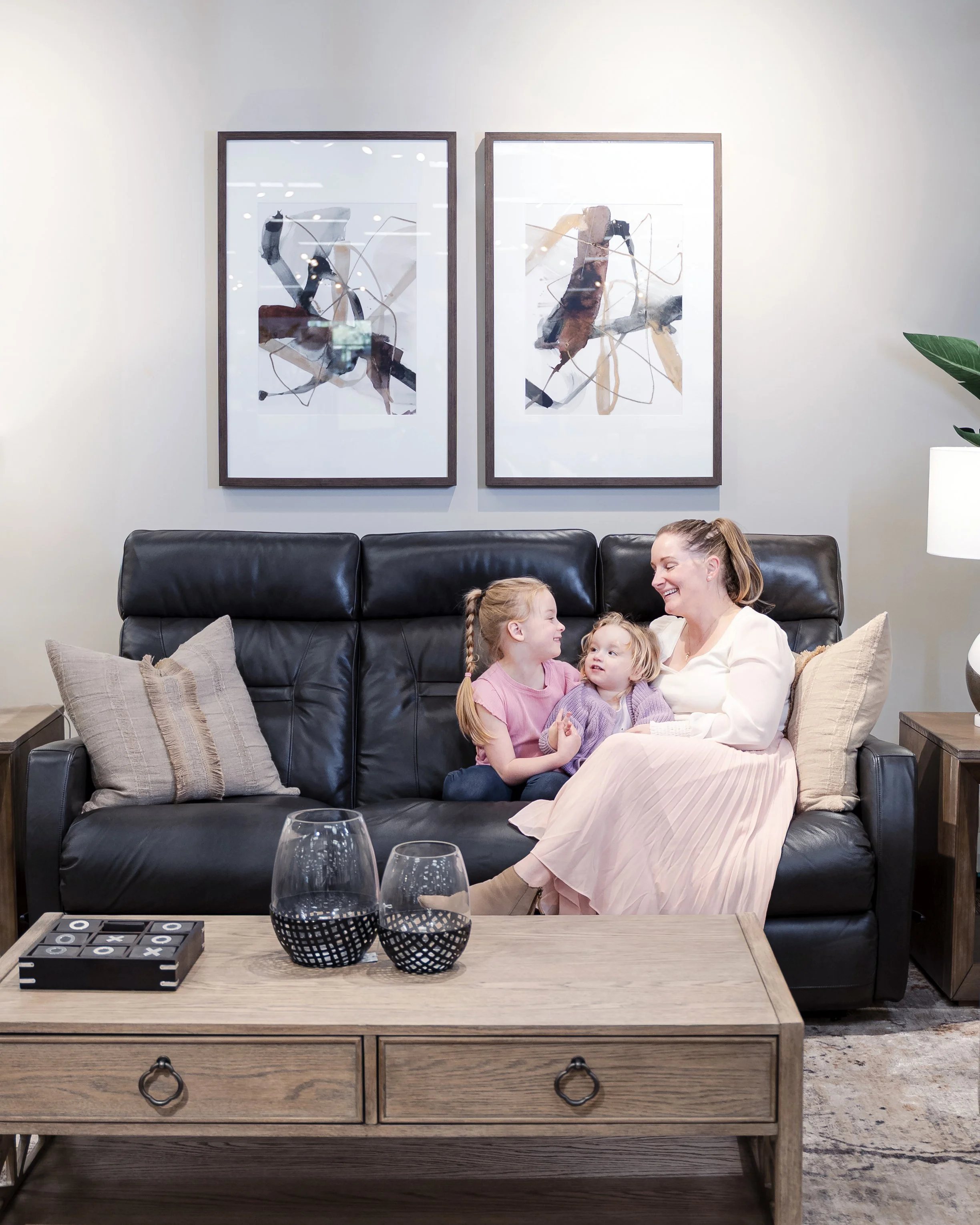 A woman and two young girls sitting on a black leather sofa in a living room, smiling and playing together, with abstract artwork hanging on the wall behind them.