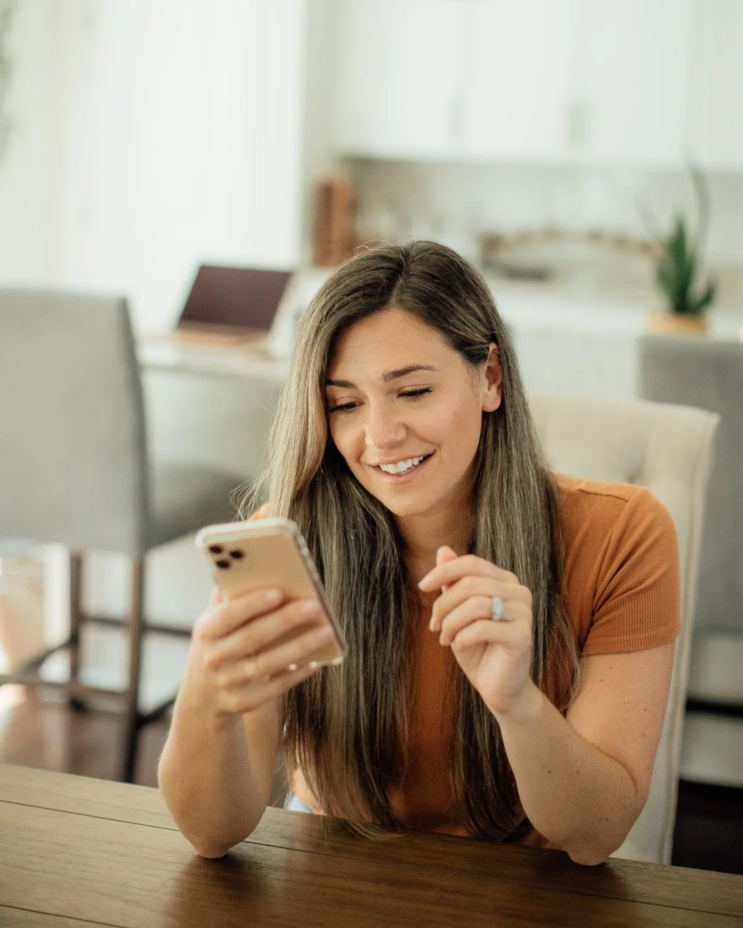 A woman with long brown hair, wearing an orange shirt, smiling while looking at her cellphone in a bright kitchen or dining area.