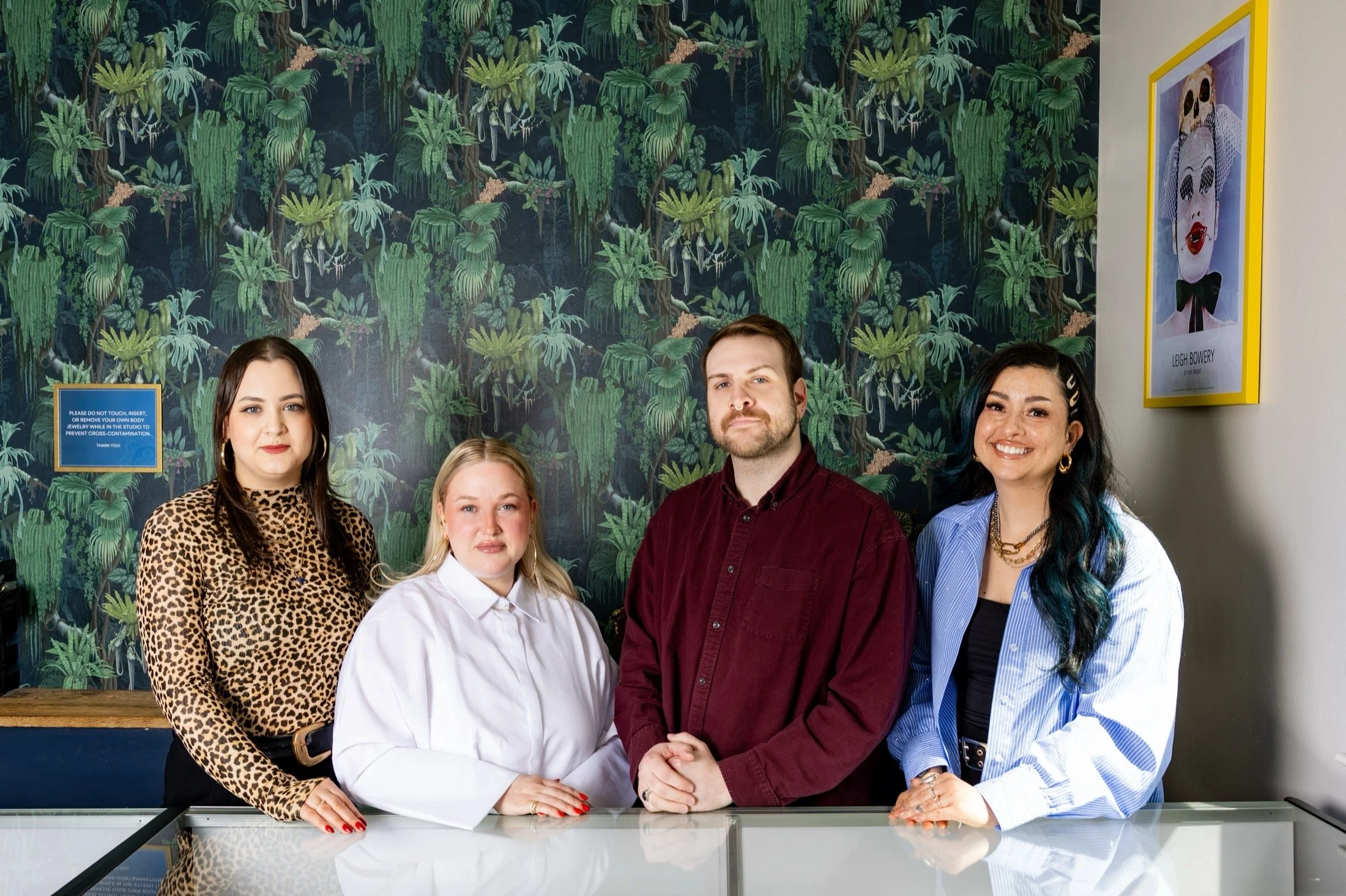Four smiling individuals standing behind a jewelry display case in a store, with a leafy, nature-themed wallpaper in the background.