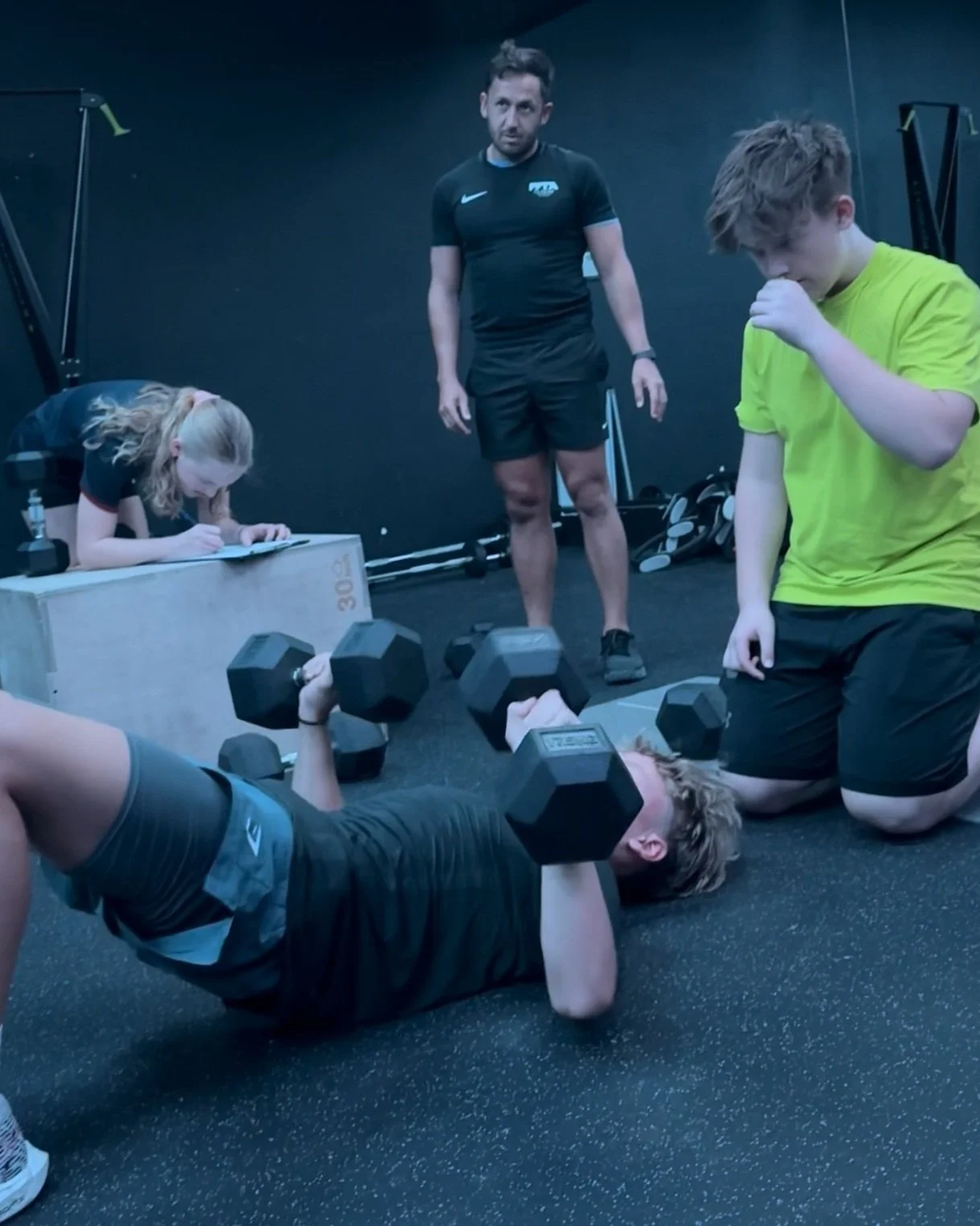 A woman is lying on her back, lifting dumbbells while a young boy kneels nearby, and a girl writes on a clipboard in a gym setting with fitness equipment in the background.