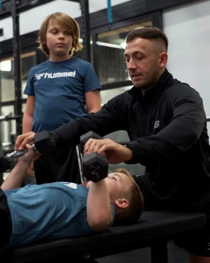 A young boy is lying on a workout bench performing a dumbbell chest press, while an adult man assists him, with a girl standing nearby watching in a gym setting.