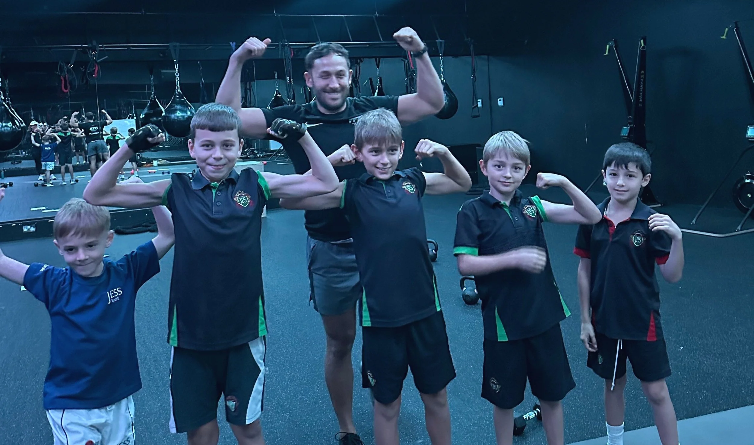 Group of five boys and one adult man in a gym posing with flexed biceps, fitness equipment in the background.