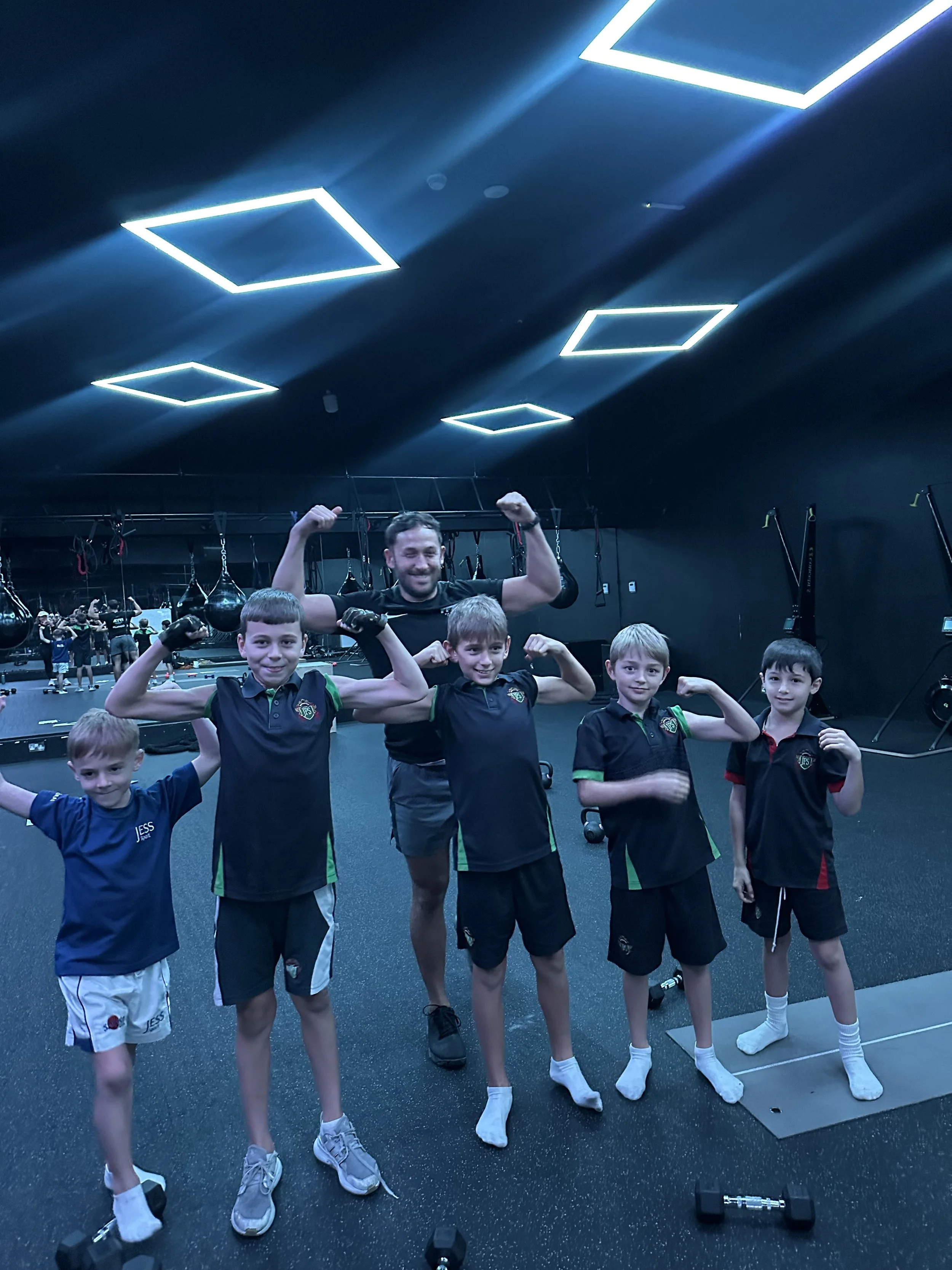 Group of five young boys and a man in a gym, flexing their muscles and smiling. The boys are wearing sports uniforms and the man is shirtless. The gym has black walls and modern rectangular ceiling lights.