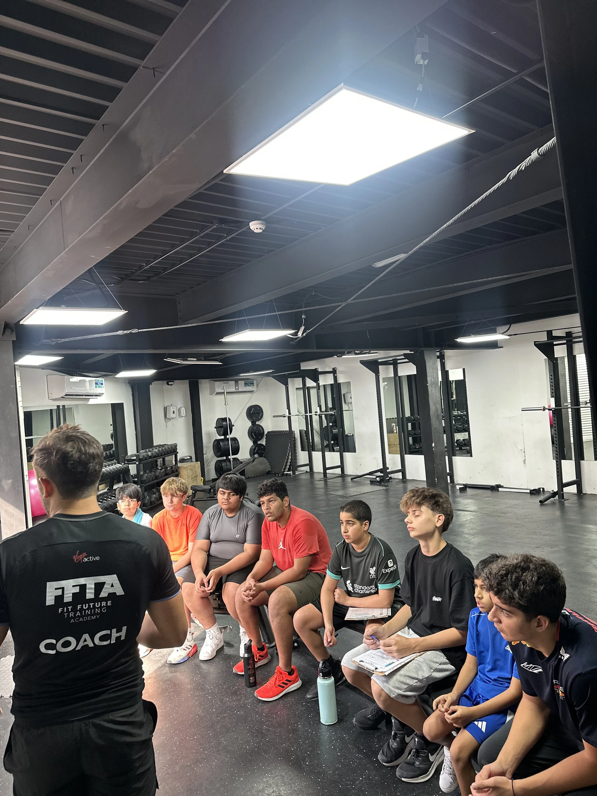 A coach is speaking to a group of young athletes seated on gym benches in a fitness training facility, with workout equipment visible in the background.