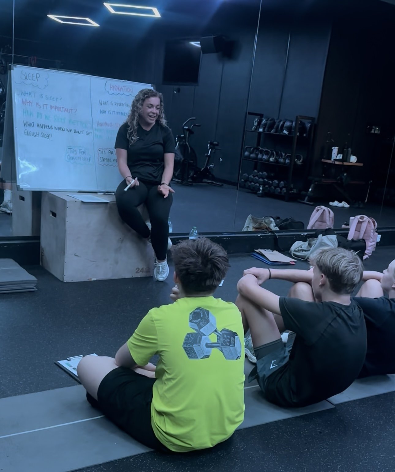 A young woman giving a fitness or health class to a group of children, sitting on the floor in a gym with workout equipment in the background, whiteboard with notes about sleep and hydration, and children attentively listening.