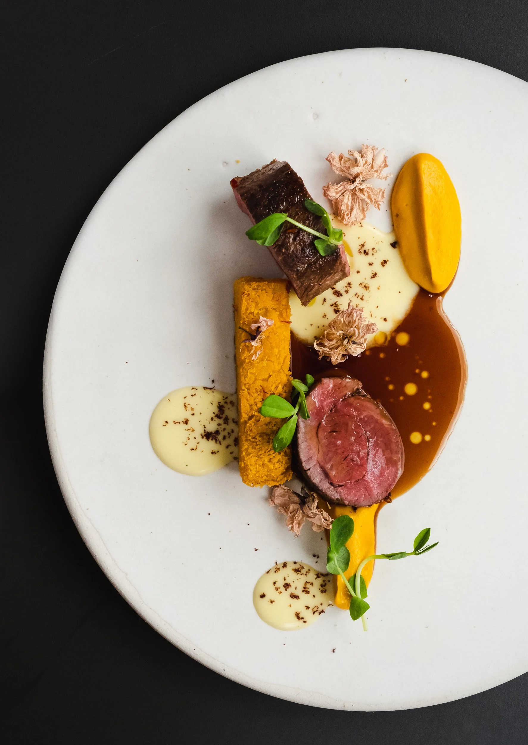Elegant plated dish featuring a slice of seared steak, a yellow cylindrical croquette, a piece of tender venison, garnished with herbs and edible flowers, with sauces and purées on a white plate against a black background.