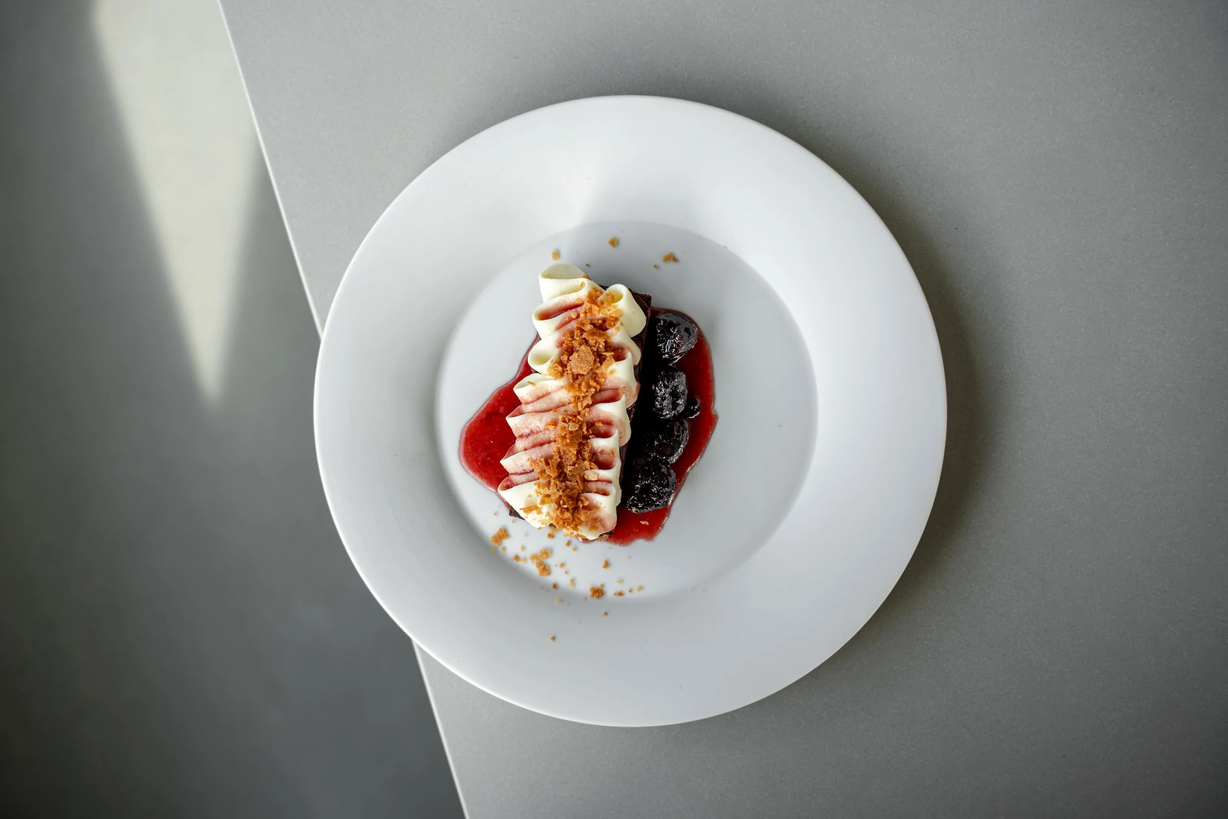 A slice of cheesecake topped with crumbled cookies and drizzled with strawberry and chocolate sauces, served on a white plate with a fruit compote underneath.