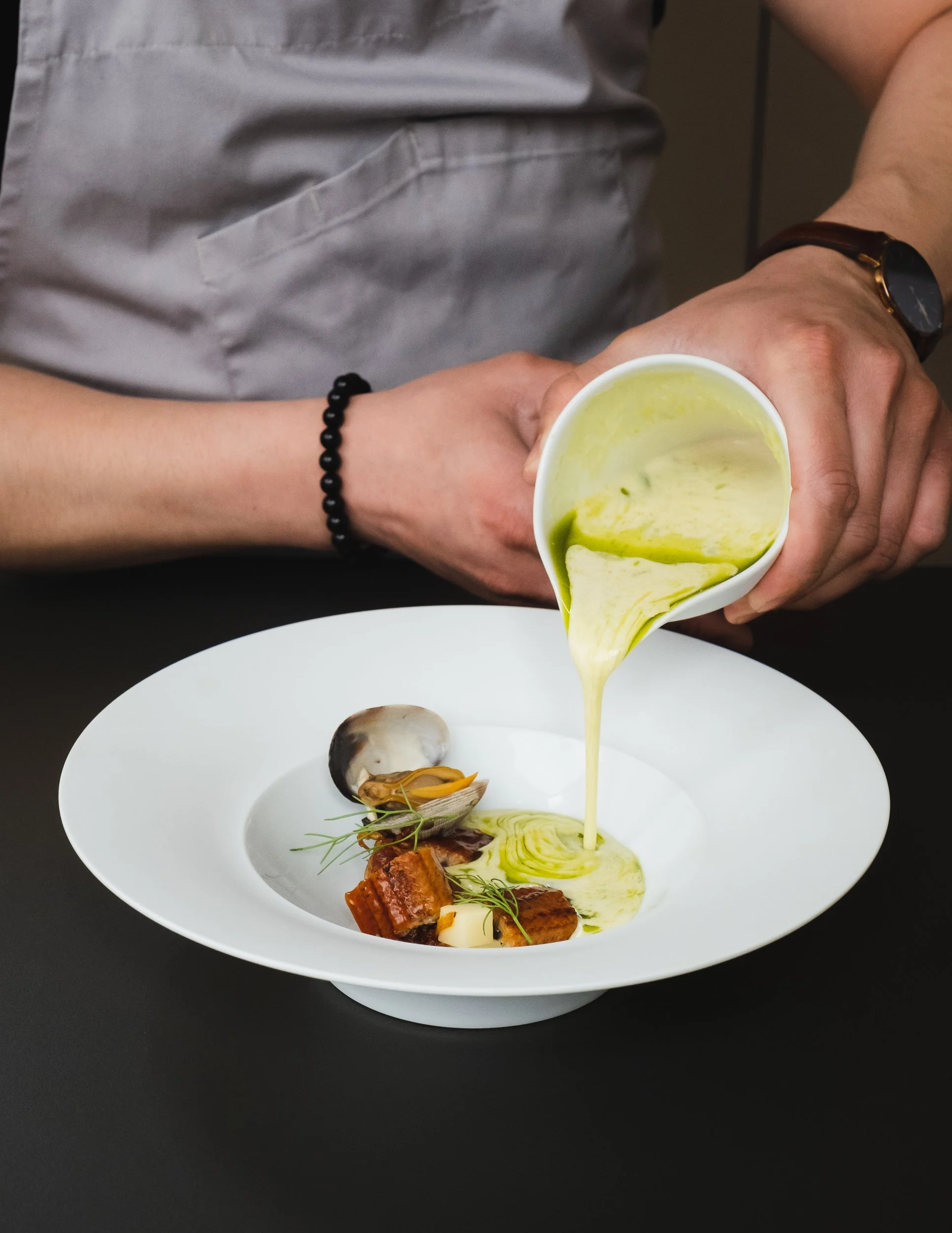 Chef pouring green sauce over gourmet seafood dish with shellfish and garnishes on a white plate.