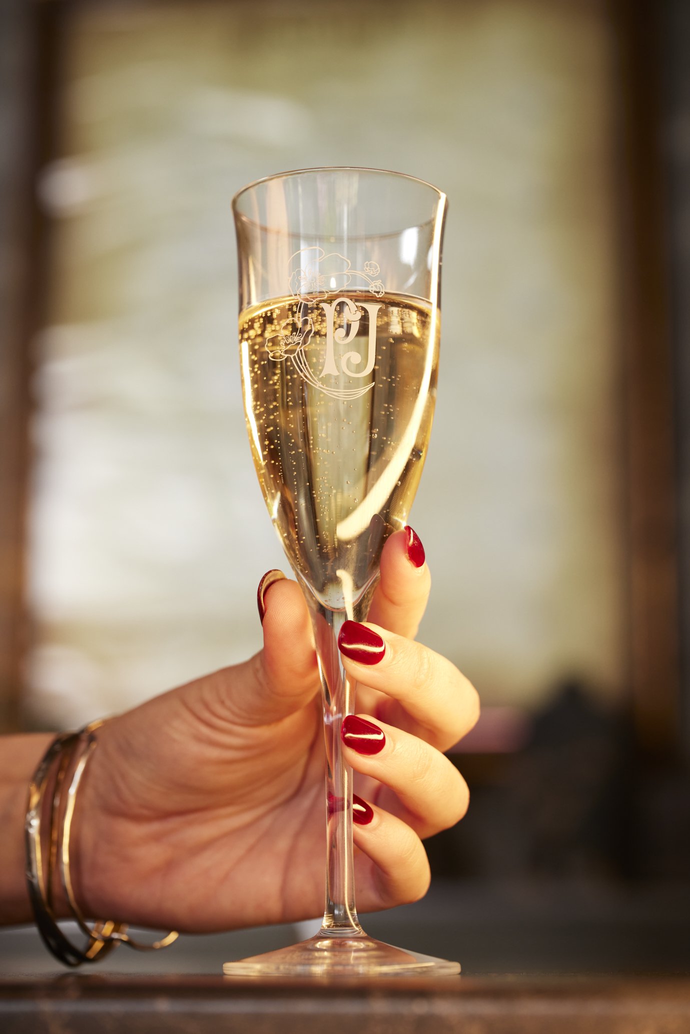 A woman with red nail polish and gold bracelets holding a flute of sparkling wine or champagne indoors with a blurred background.