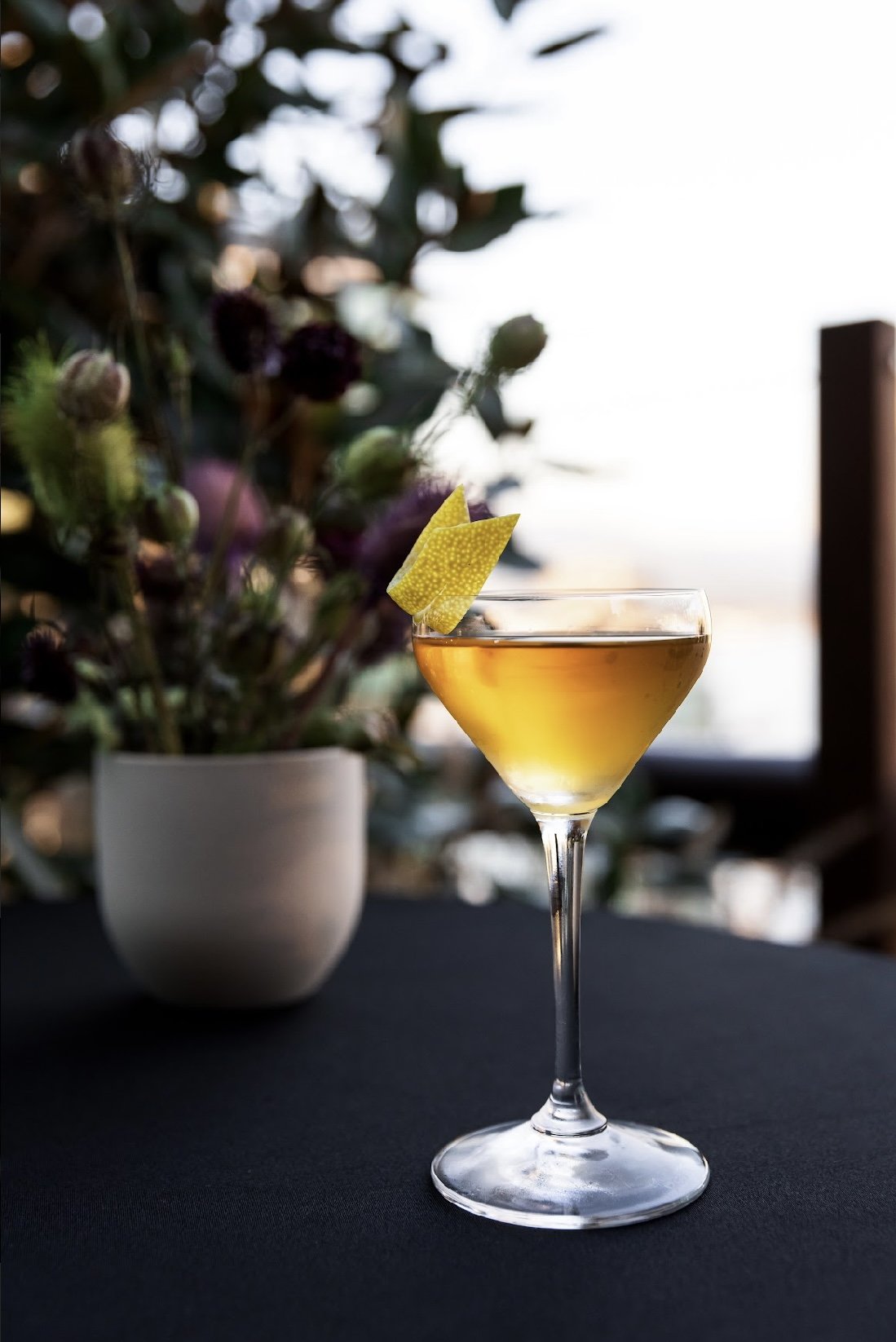 A yellow cocktail with lemon garnish in a cocktail glass on a dark table, with a vase of flowers in the background.
