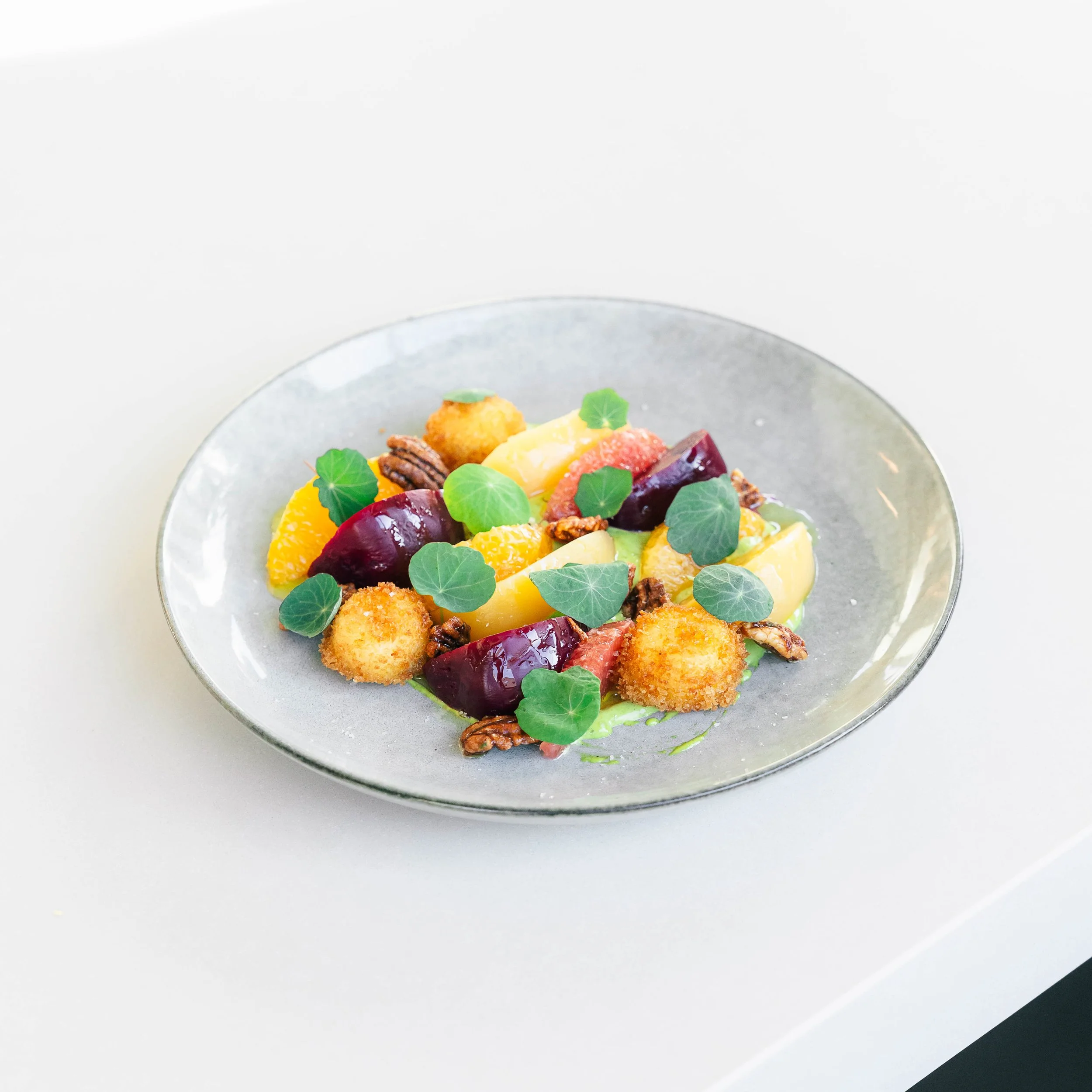 A plated salad with roasted beets, orange segments, microgreens, toasted pecans, and breaded cheese balls on a gray dish.