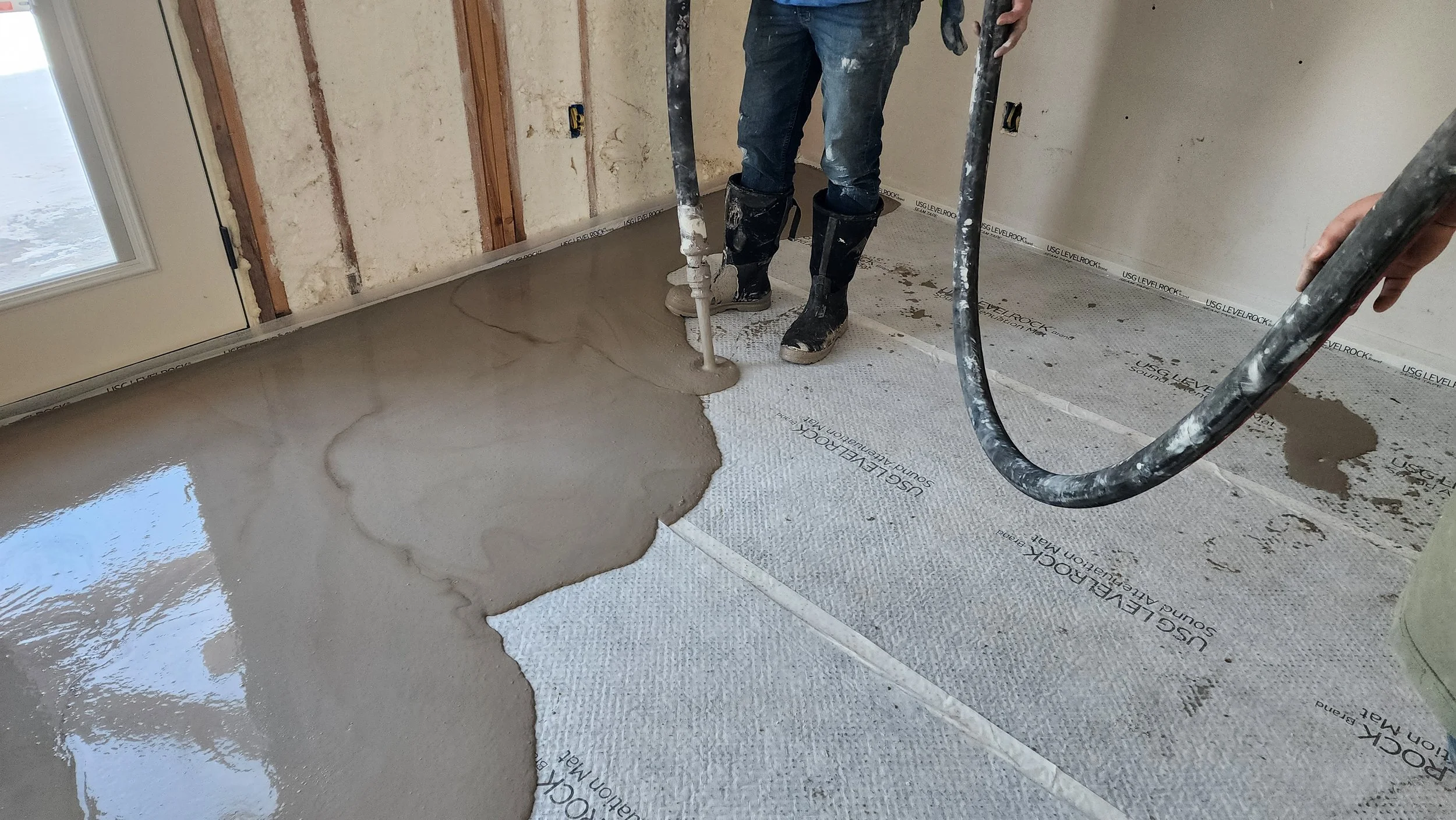 Person pouring wet concrete onto a floor in a room under construction, with drywall and flooring materials visible.