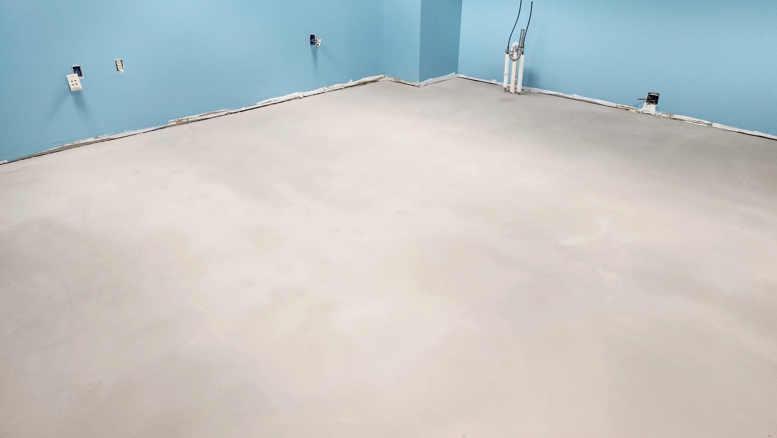 Empty room with blue walls and a light-colored floor, with electrical outlets and plumbing fixtures visible.