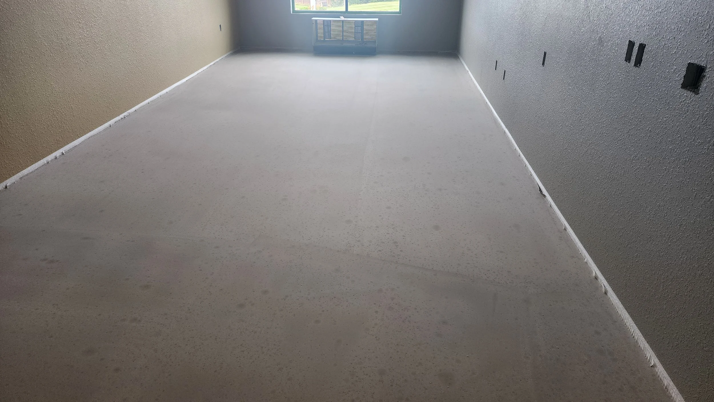 An empty room with beige and gray walls, a window at the far end, and a light-colored floor with visible seams.