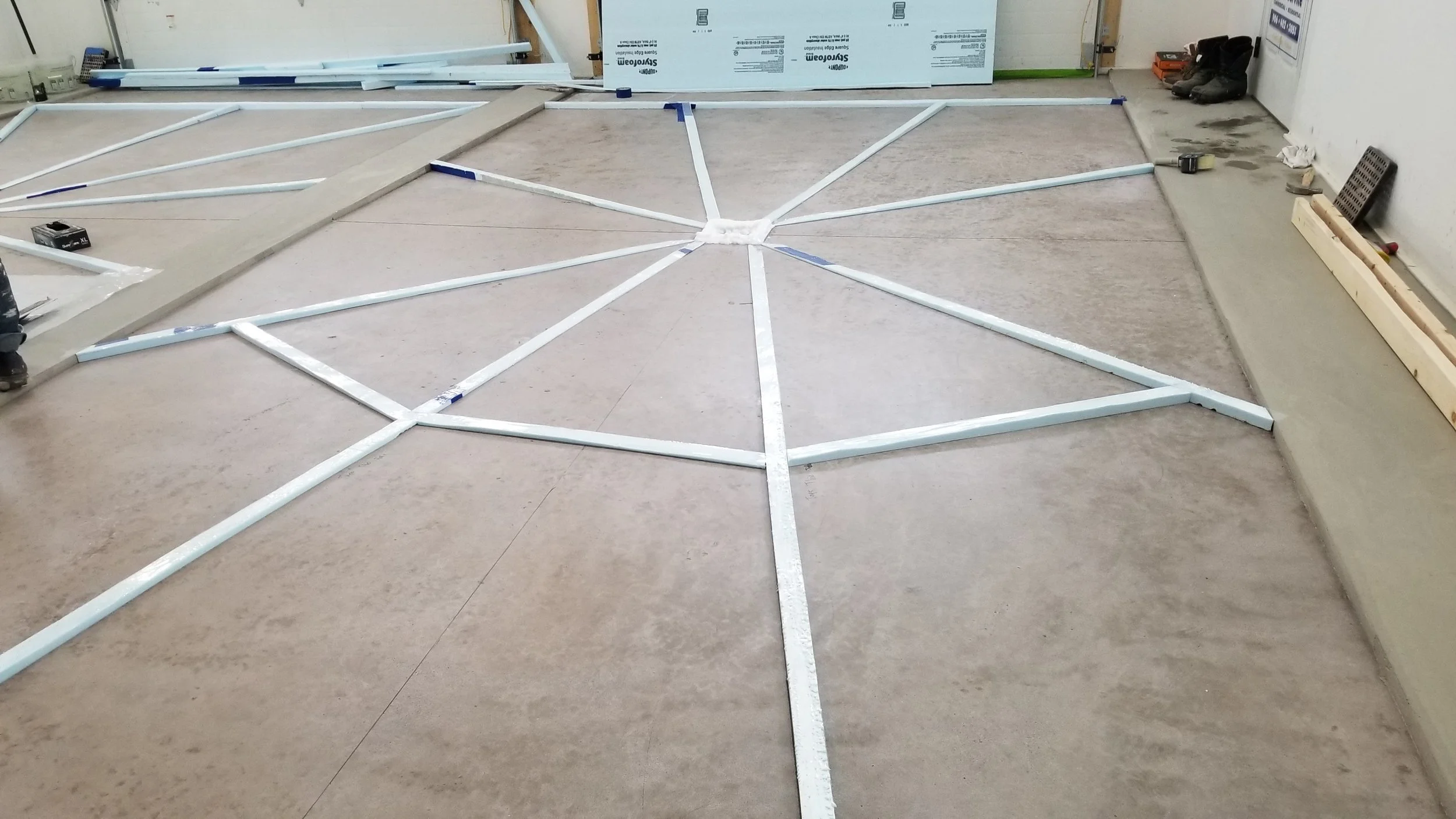 Construction site with white metal framing laid out on the floor forming geometric shapes, tools and equipment scattered around, and a partially constructed wall in the background.