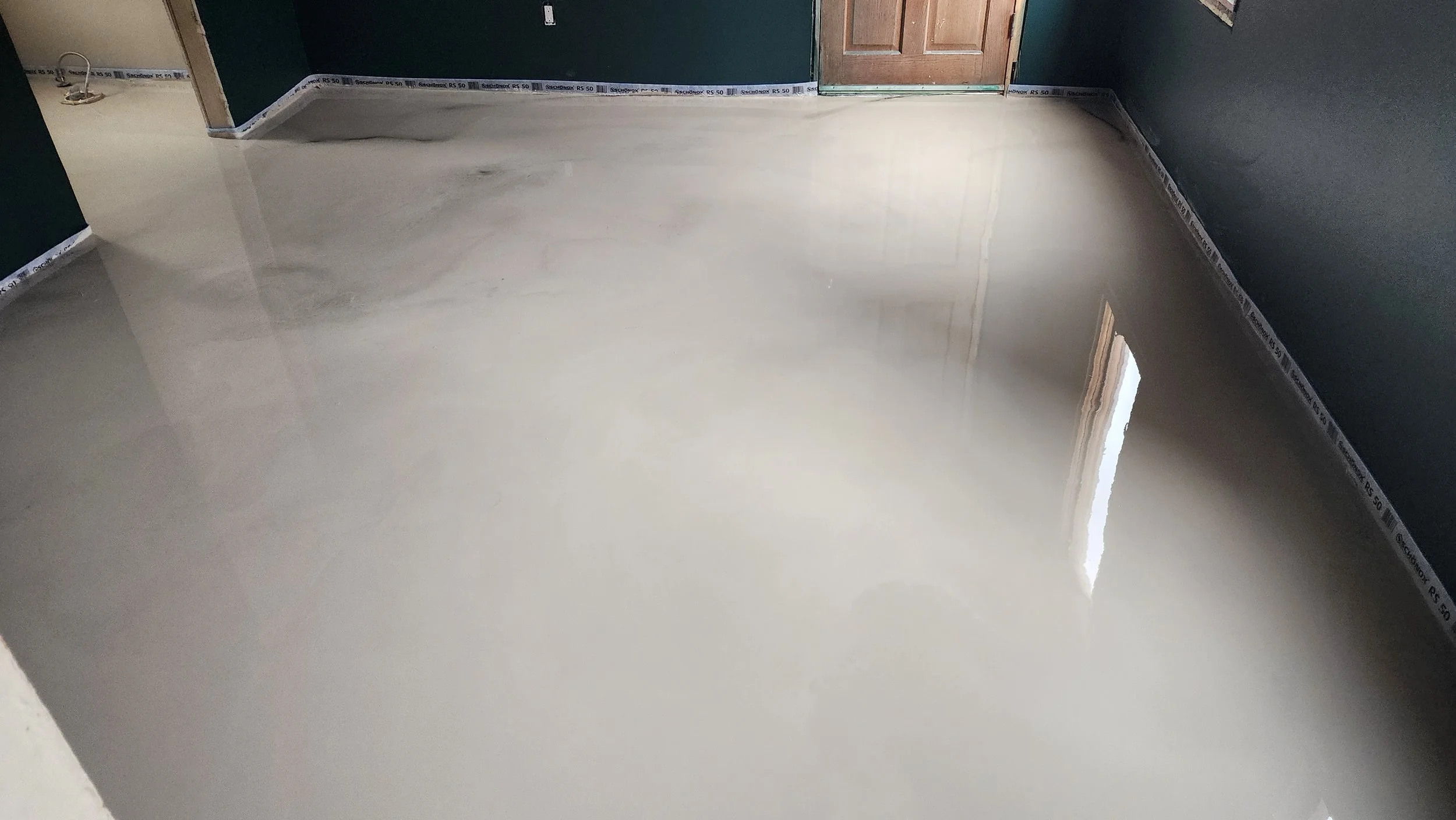 Freshly poured concrete floor in a room with green walls and a wooden door, reflecting light from a window.